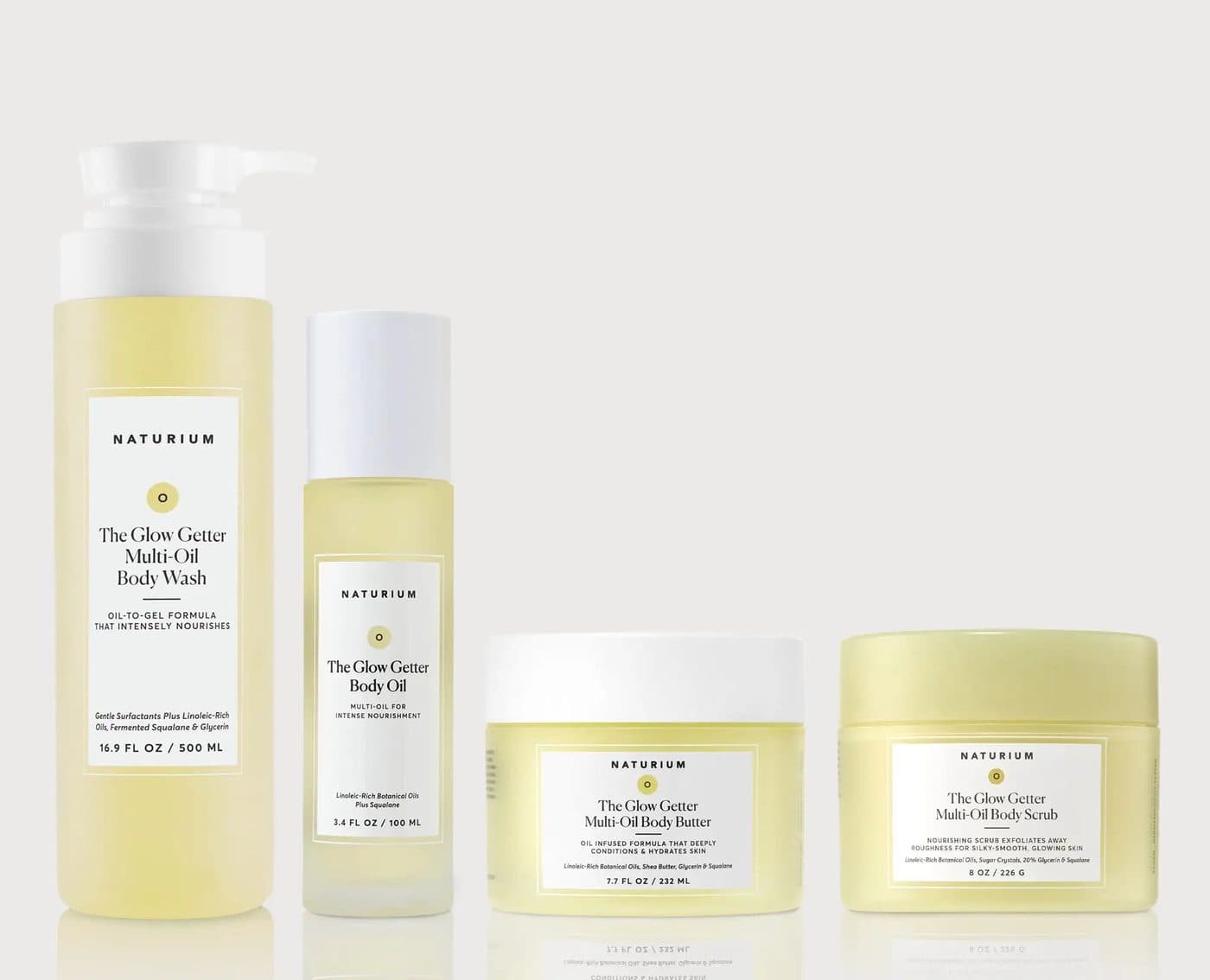 Naturium's Glow Getter body wash, body oil, body butter and body scrub in a row on a bone-white surface.