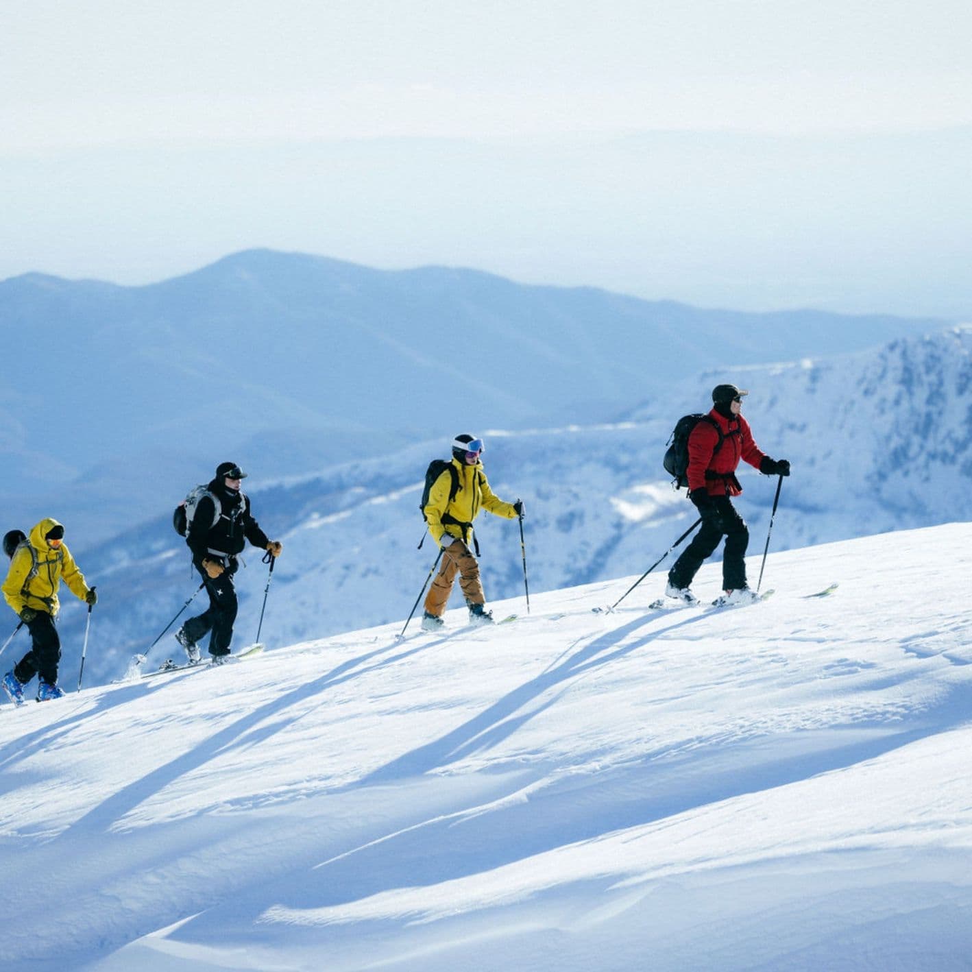 Four ski tourers in bright jackets climbing a sunlit snow ridge with mountains behind