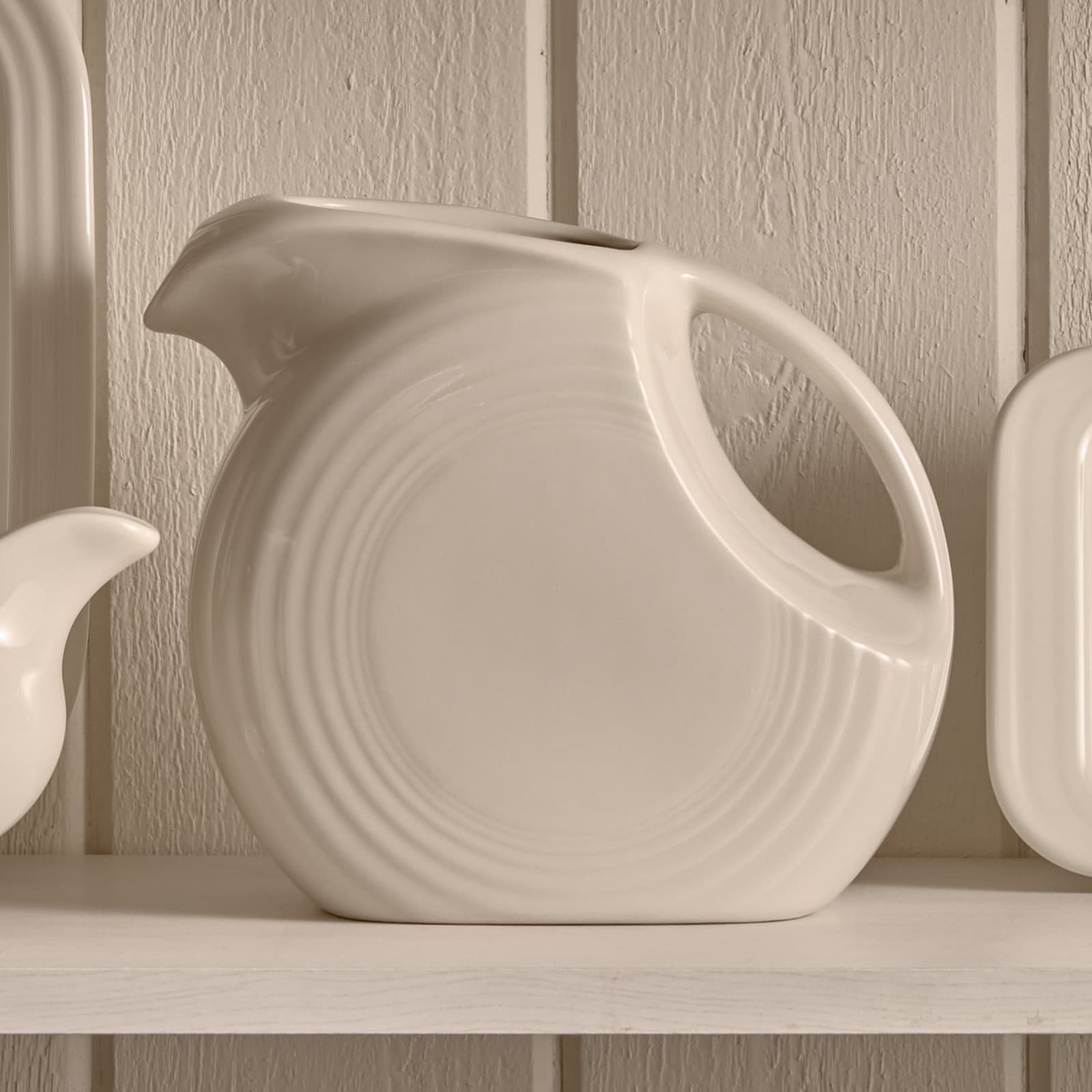 A cream ceramic disk pitcher on a white shelf against cream board paneling