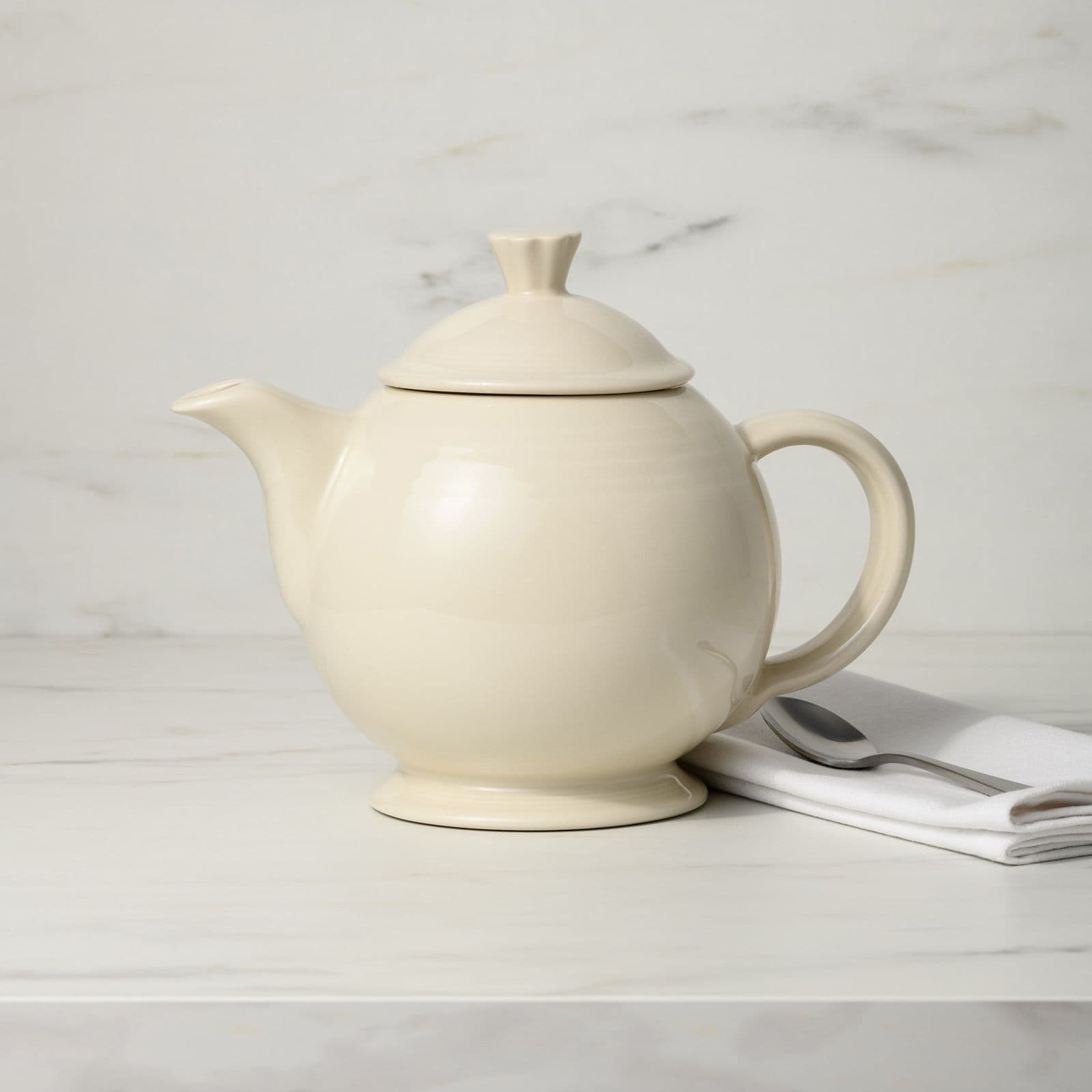 A cream ceramic teapot on a white marble counter beside a folded napkin and a spoon