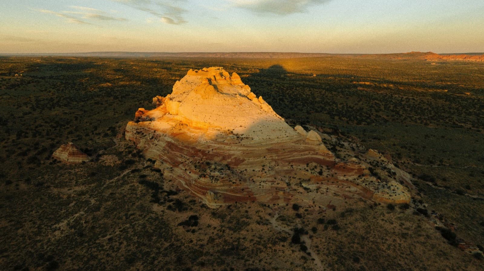 An aerial view of a banded sandstone butte at golden hour in a dark desert plain