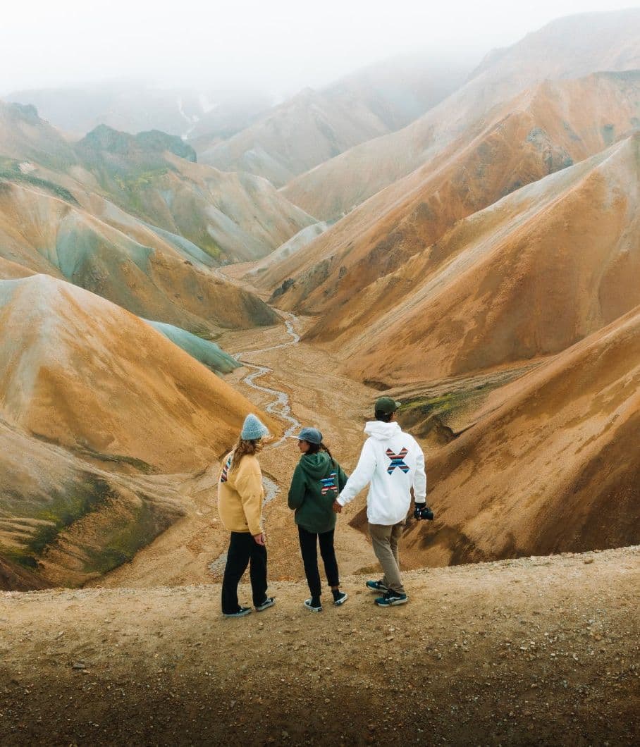 Three hikers seen from behind on a trail below striped orange and green mountains under low cloud