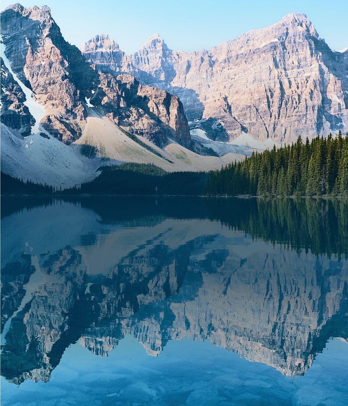 Snow streaked mountain peaks mirrored in a still turquoise lake below a pine forest