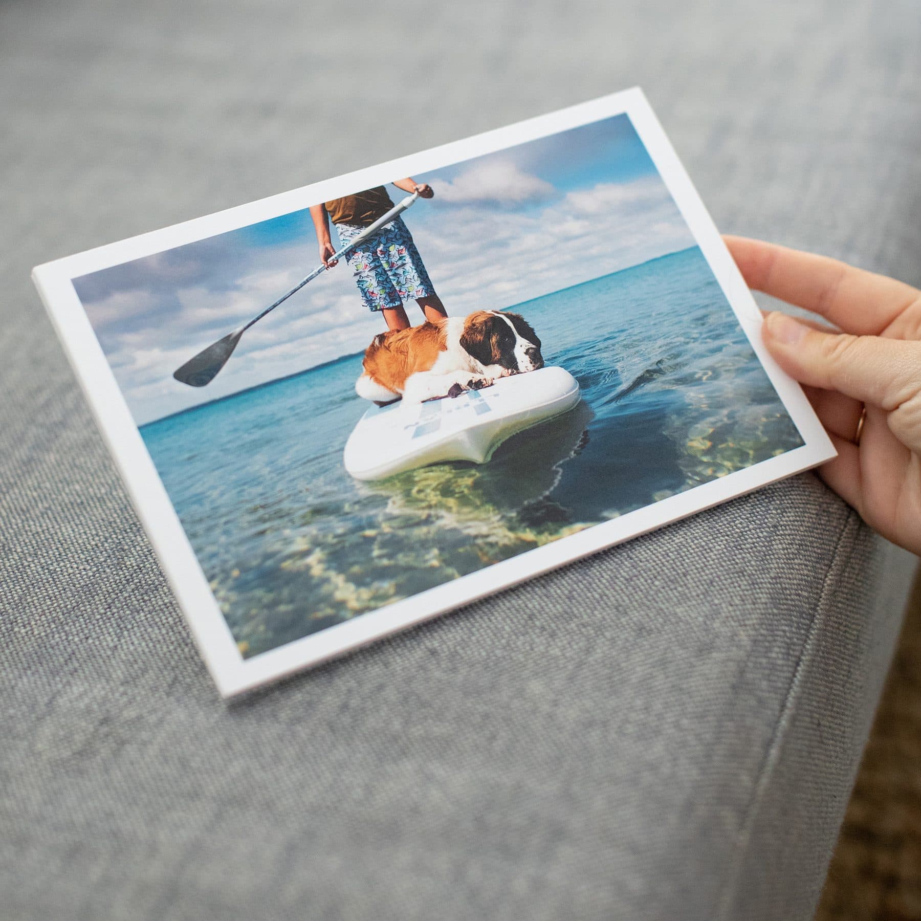 A hand holding an Mpix photo print of a person paddleboarding with a large dog on clear blue water, above a grey sofa.