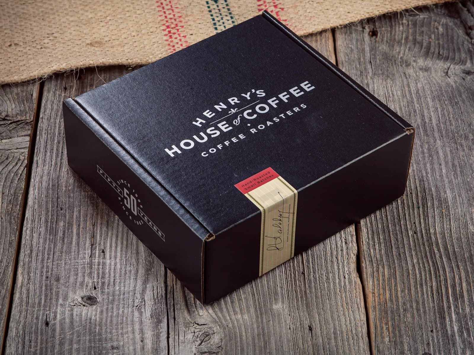 A black Henry's House of Coffee gift box on weathered wooden boards beside a burlap coffee sack.