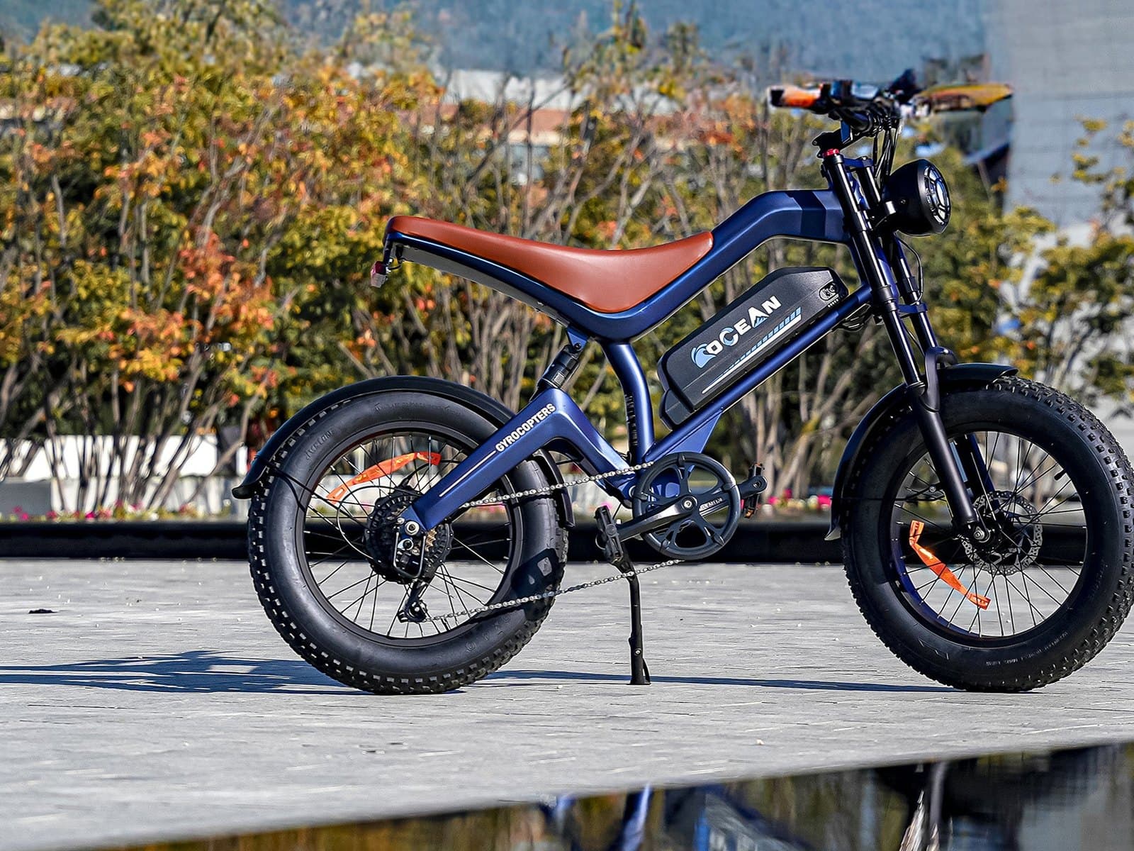 A navy fat-tire e-bike with a cognac saddle parked side on on a pale stone plaza in low sun, autumn trees behind.