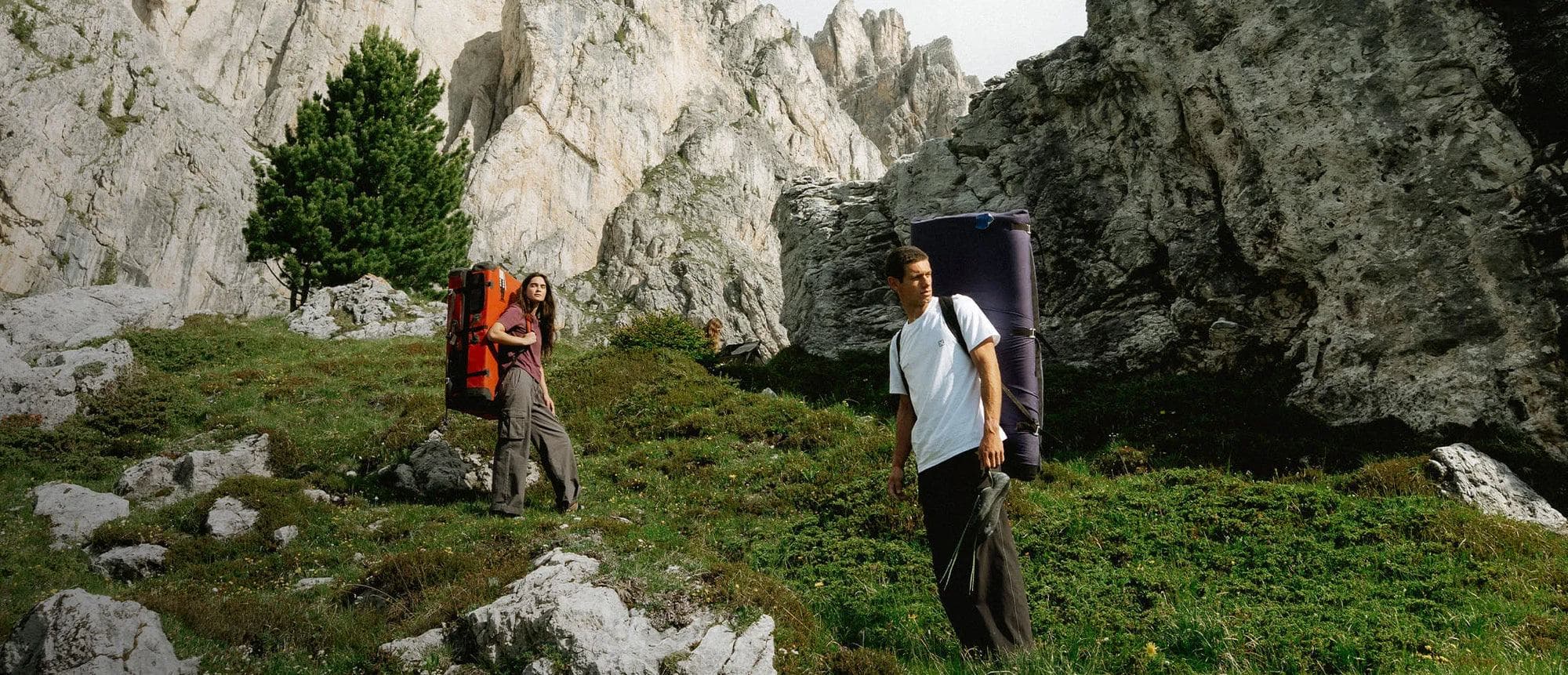 Two climbers carrying bouldering crash pads across a grassy slope below pale limestone cliffs.