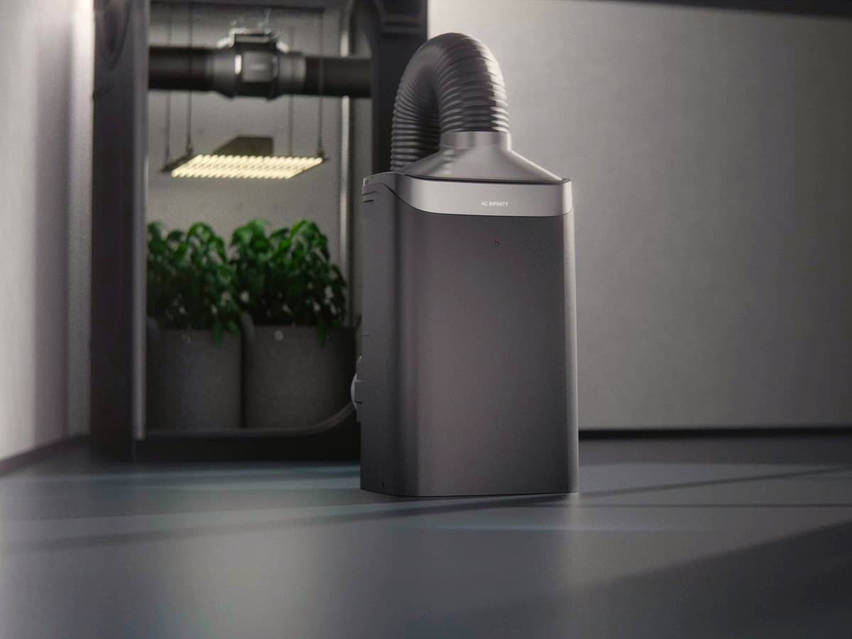 A dark grey AC Infinity unit with a flexible duct standing beside an open grow tent of leafy plants under a light bar.