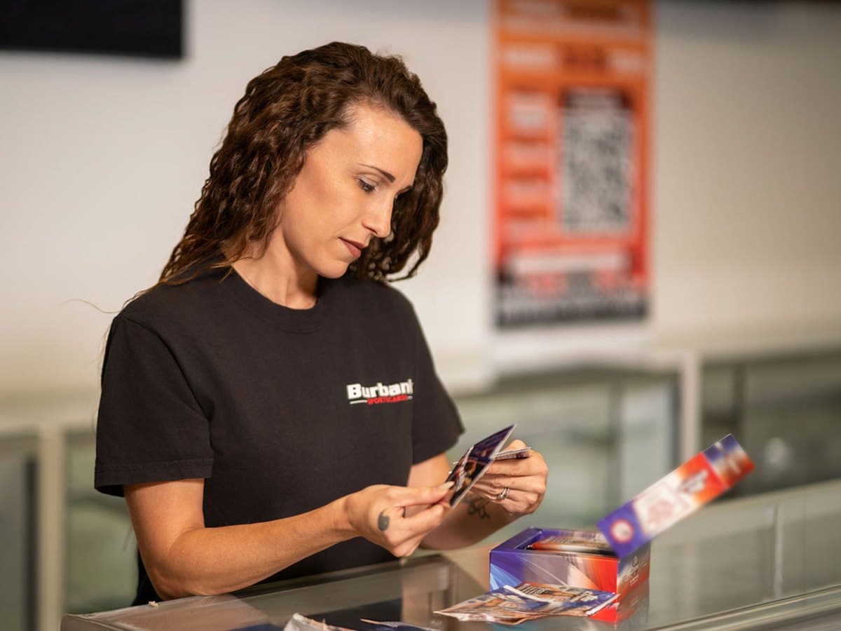 A woman in a black Burbank Sportscards T-shirt looking through freshly opened trading cards over a glass display counter.