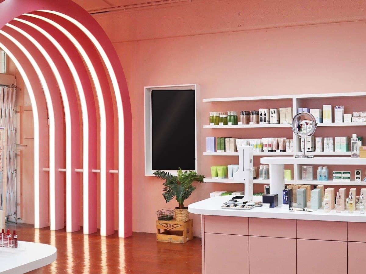 A pink beauty store with an arch of glowing white bands, shelves of skincare and a white counter beside a potted palm.