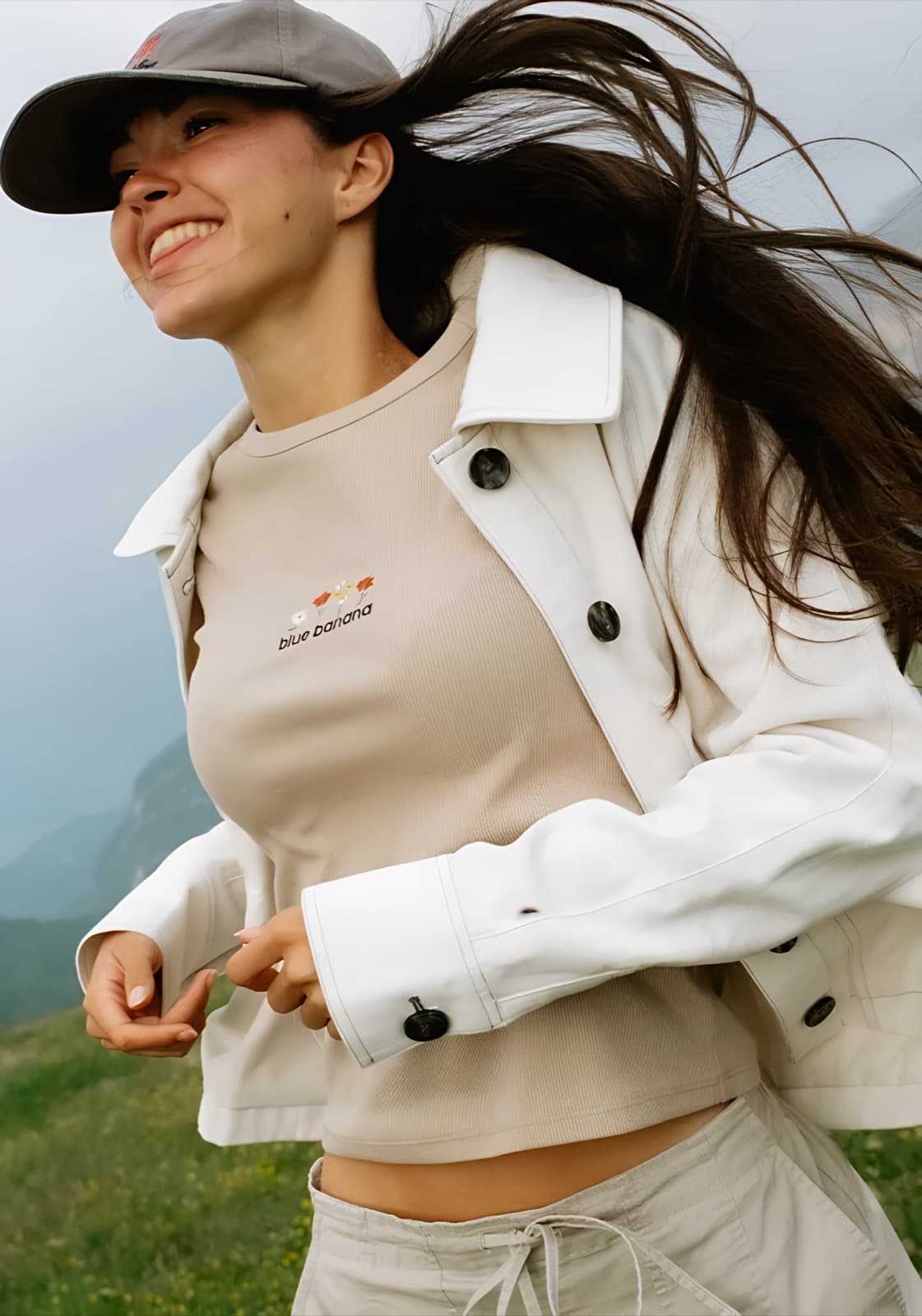 A woman in a white jacket and cap laughing in the wind on a misty green hillside.