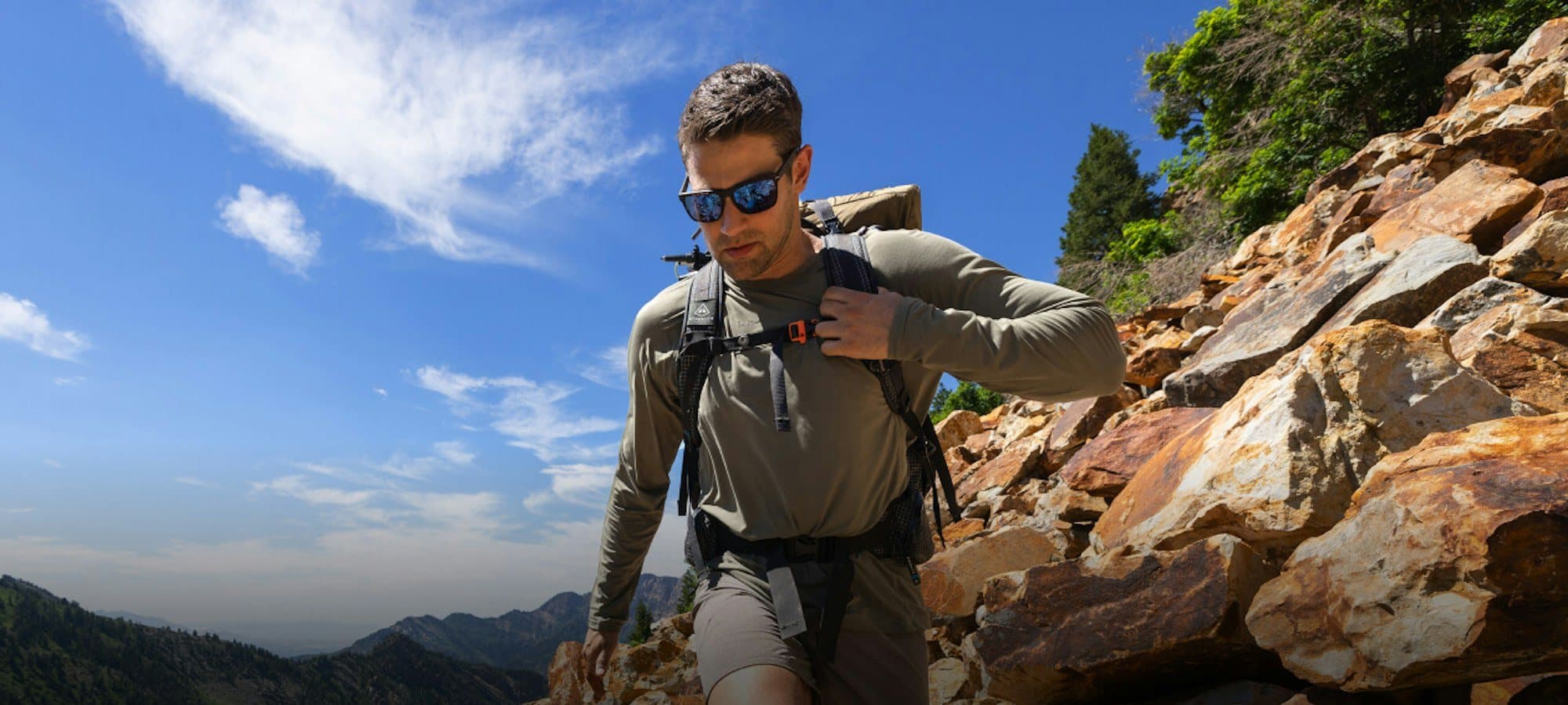 A hiker in ROKA sunglasses and a trail pack climbing a rocky ridge under a blue sky, mountains behind.