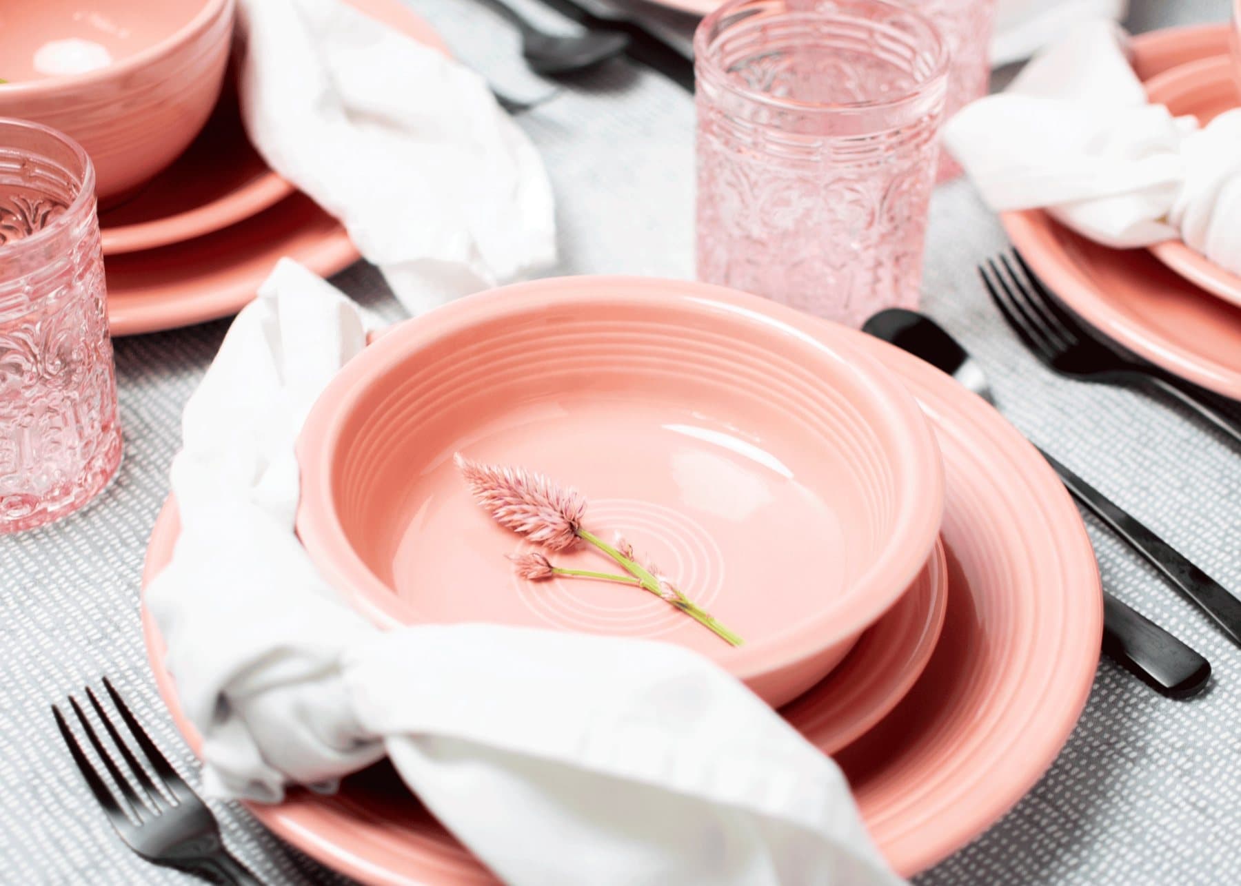 A pink Fiesta bowl on a matching plate with a white napkin and black flatware, pink pressed glasses behind, on grey woven linen.