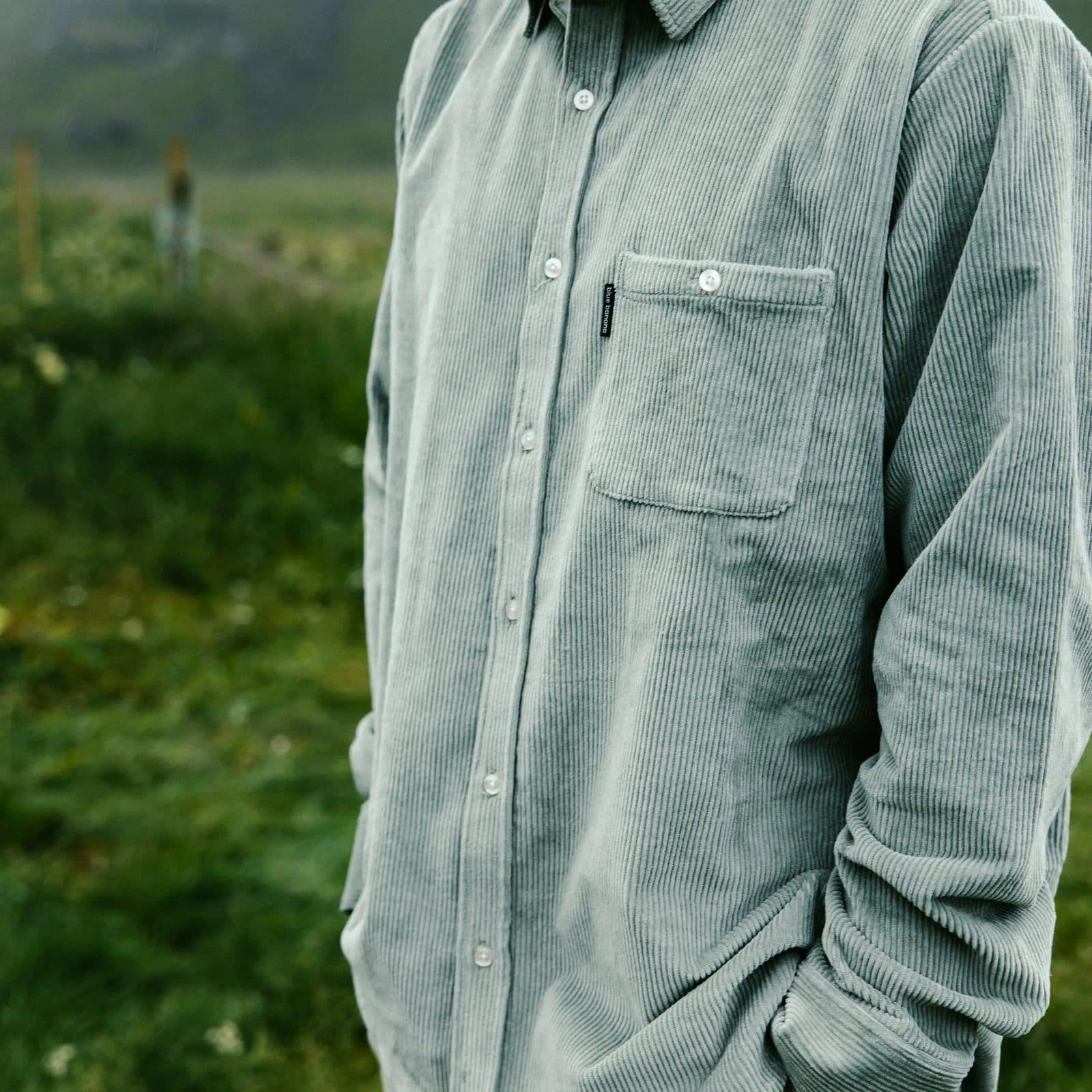 A sage green corduroy shirt with a chest pocket, worn with a hand in the trouser pocket, in a green field.