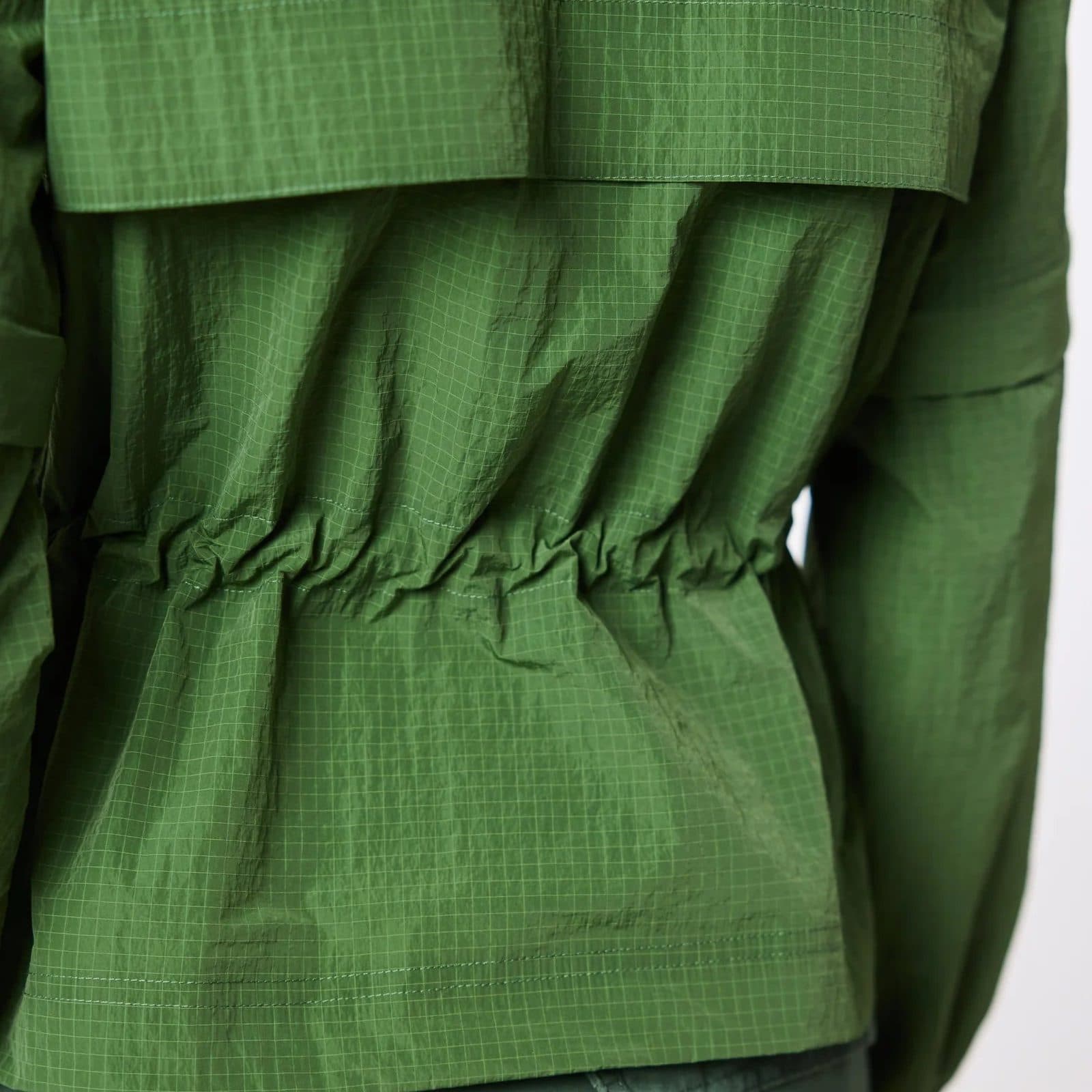 The back of a green ripstop jacket with a cinched waist, against a pale studio wall.