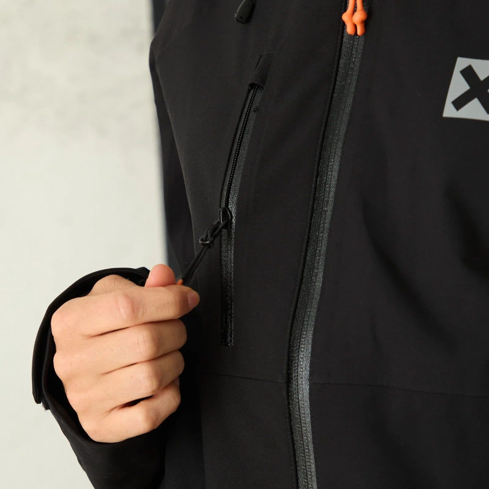 A hand pulling the chest pocket zip of a black waterproof shell jacket, orange zip pulls at the collar.