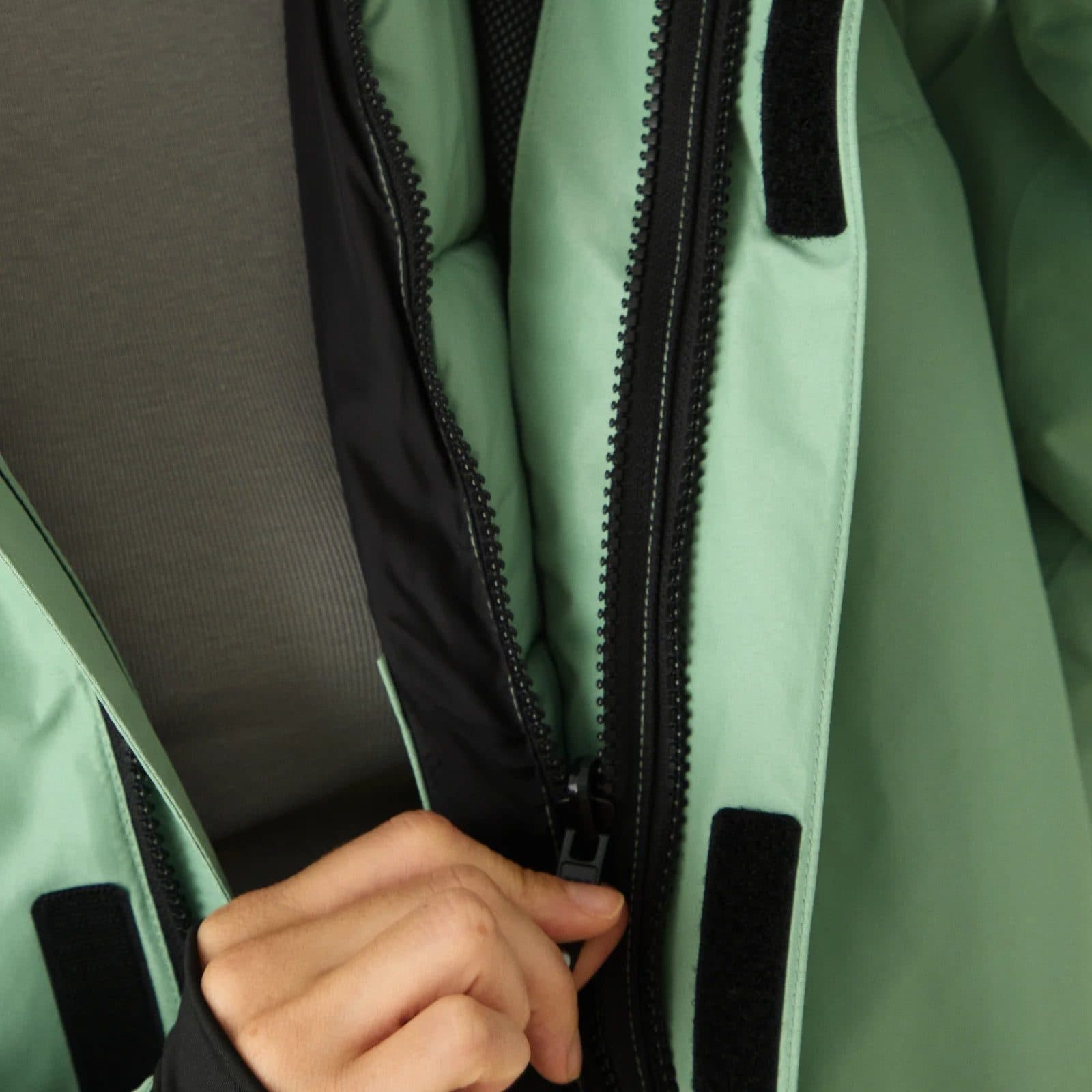 A hand holding open a mint green shell jacket at its black zip, black hook and loop tabs on the storm flap.