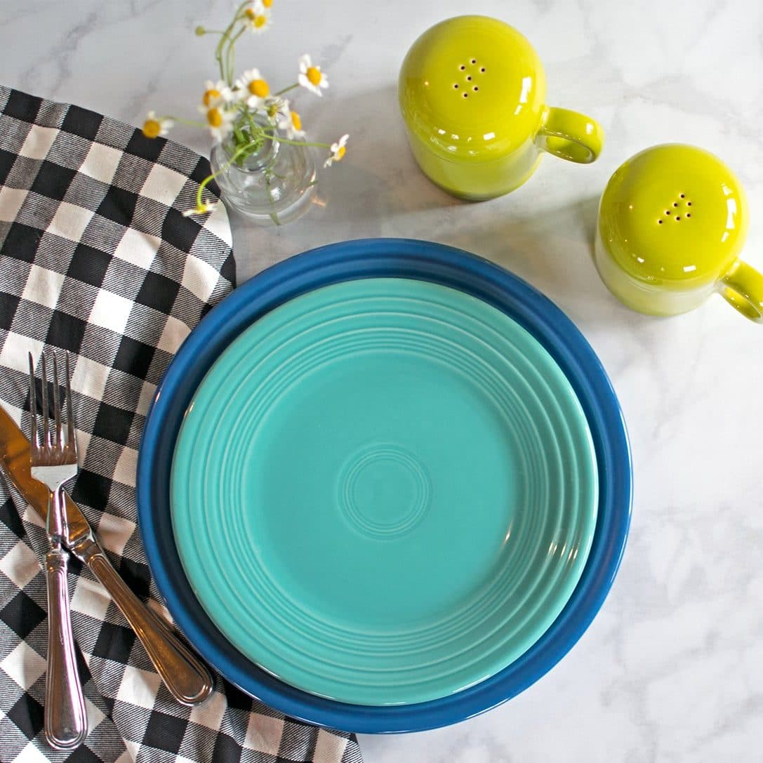 A turquoise plate on a cobalt plate beside a black gingham napkin, yellow shakers and daisies on white marble.