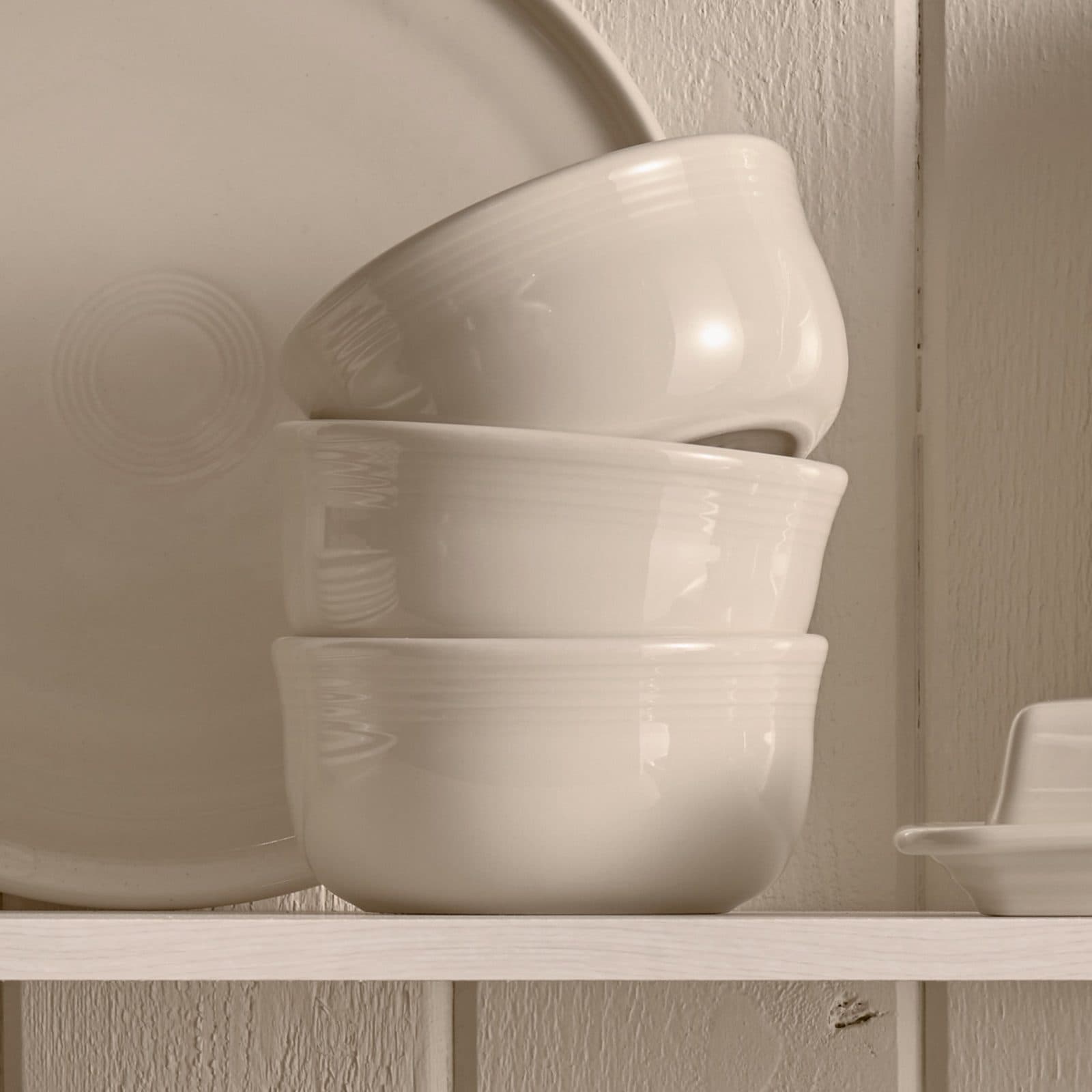 Three cream bowls stacked on a white wood shelf beside the rim of a cream plate.