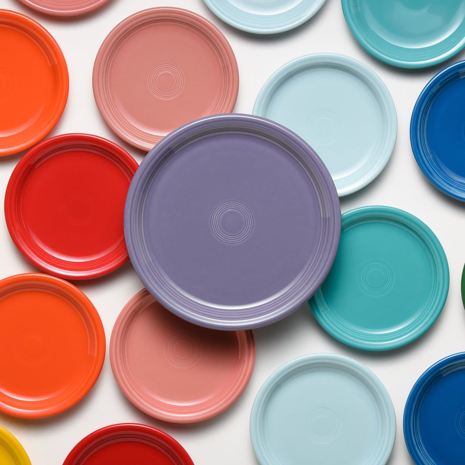 Round plates in red, coral, yellow, turquoise, sky blue and cobalt laid flat on white, one lavender plate at the centre.
