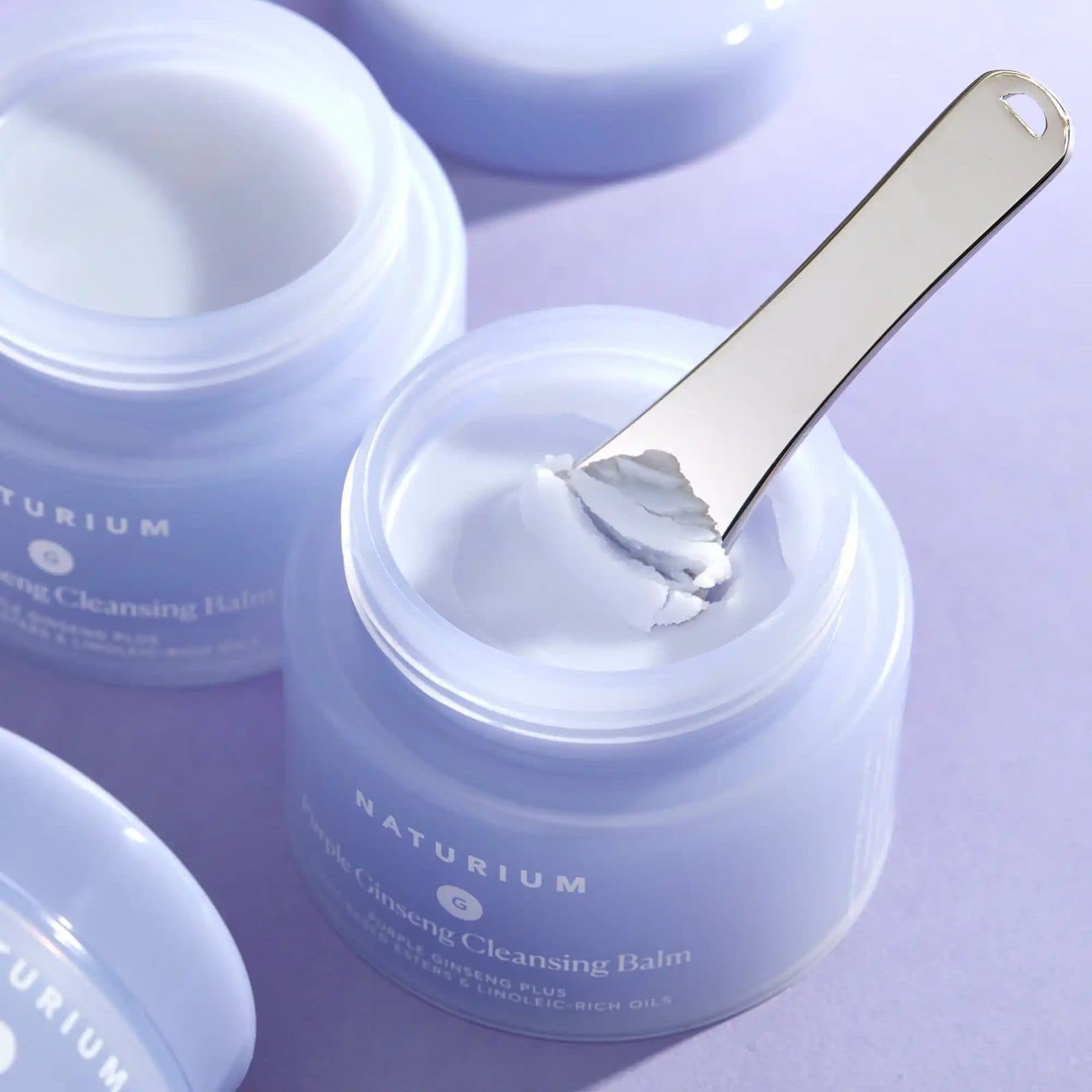 Open jars of Naturium Purple Ginseng Cleansing Balm on a lavender backdrop, a steel spatula lifting balm from one