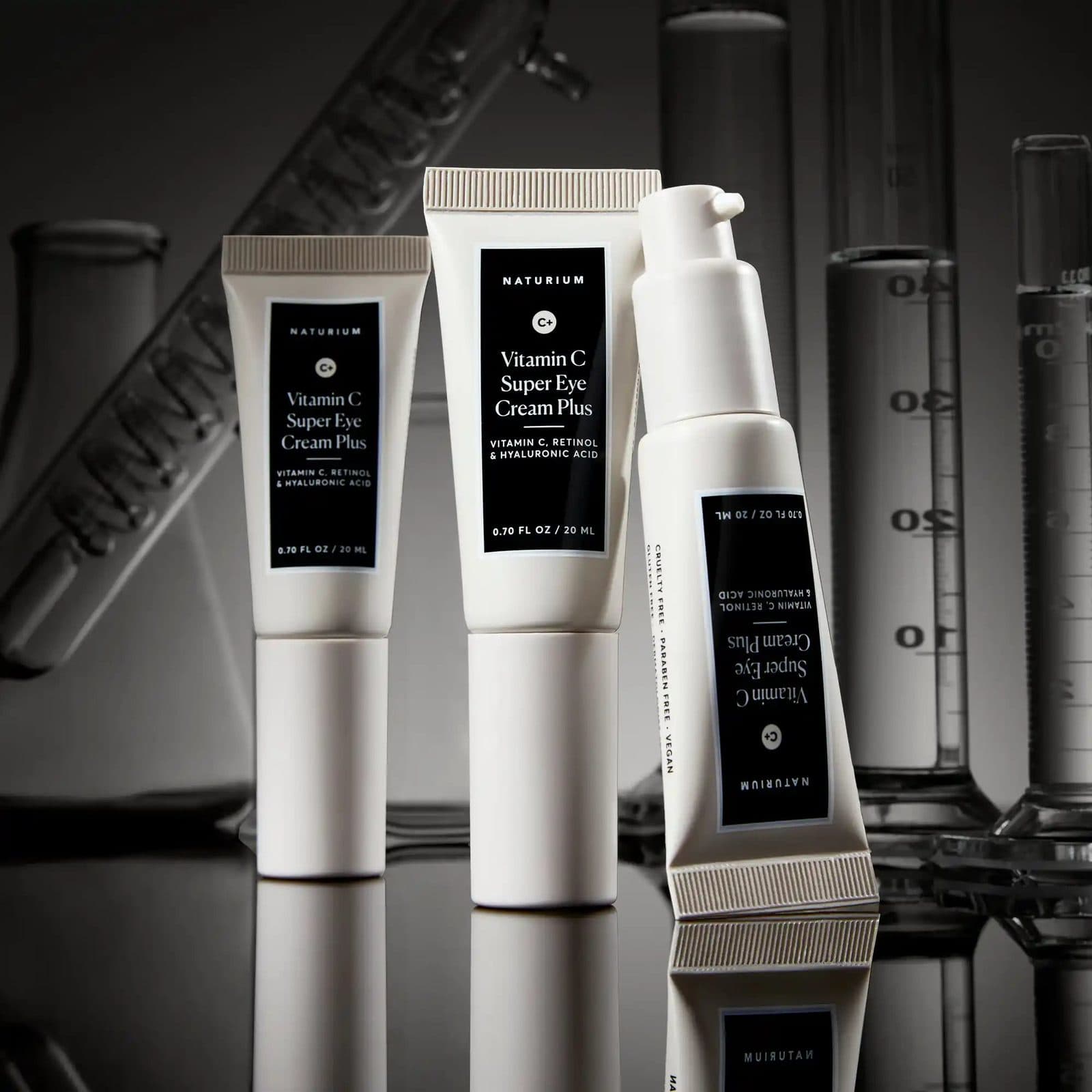 Three white eye cream tubes on a black reflective surface, with graduated cylinders and lab flasks in grey behind.