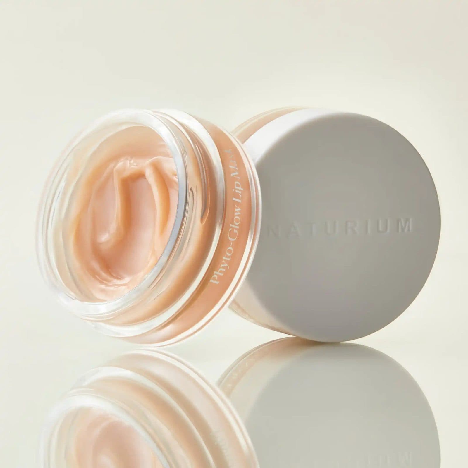 An open glass jar of peach lip balm leaning beside its pale grey lid, on a soft cream backdrop.
