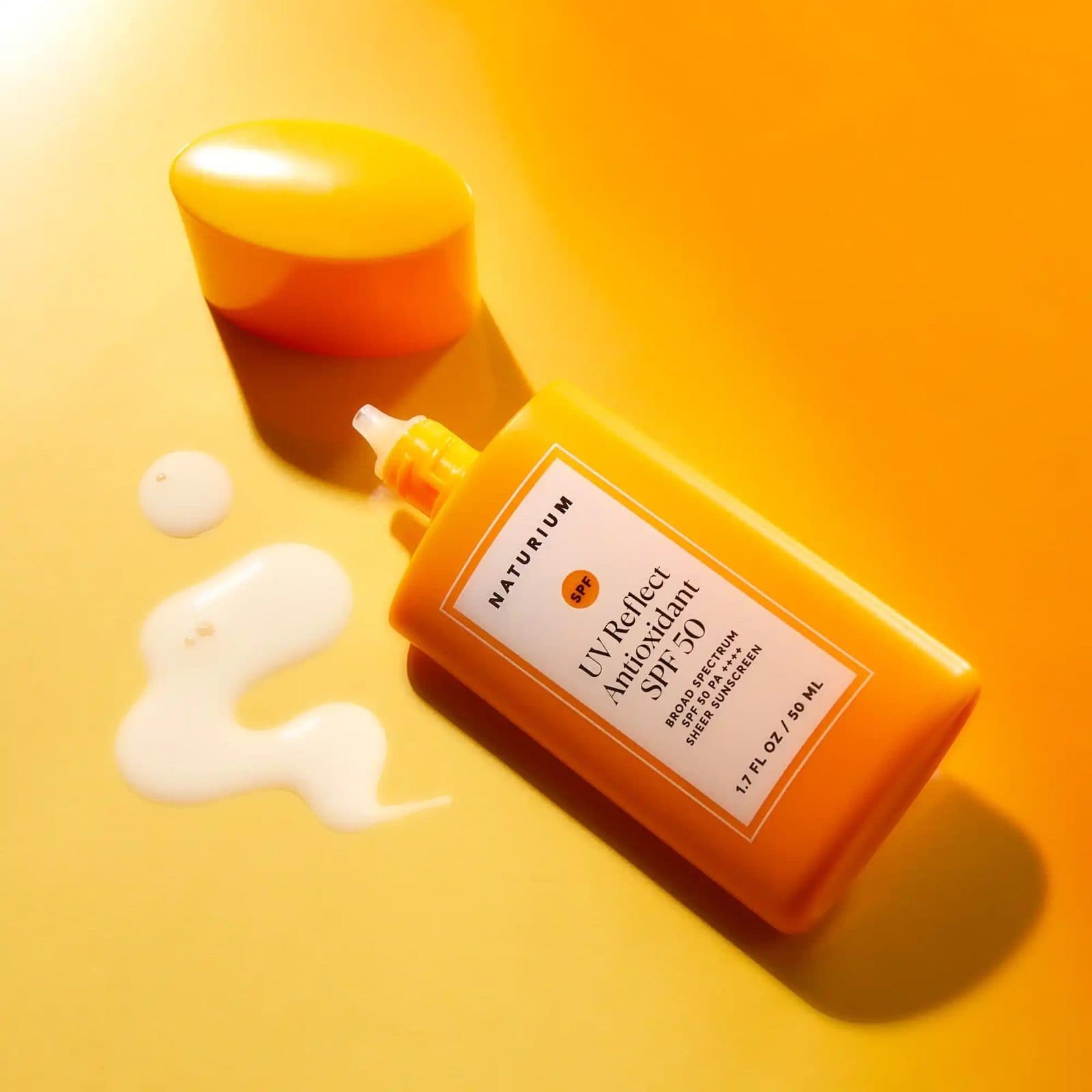 An orange bottle of SPF 50 sunscreen lying on an orange backdrop beside its cap and a swirl of lotion.