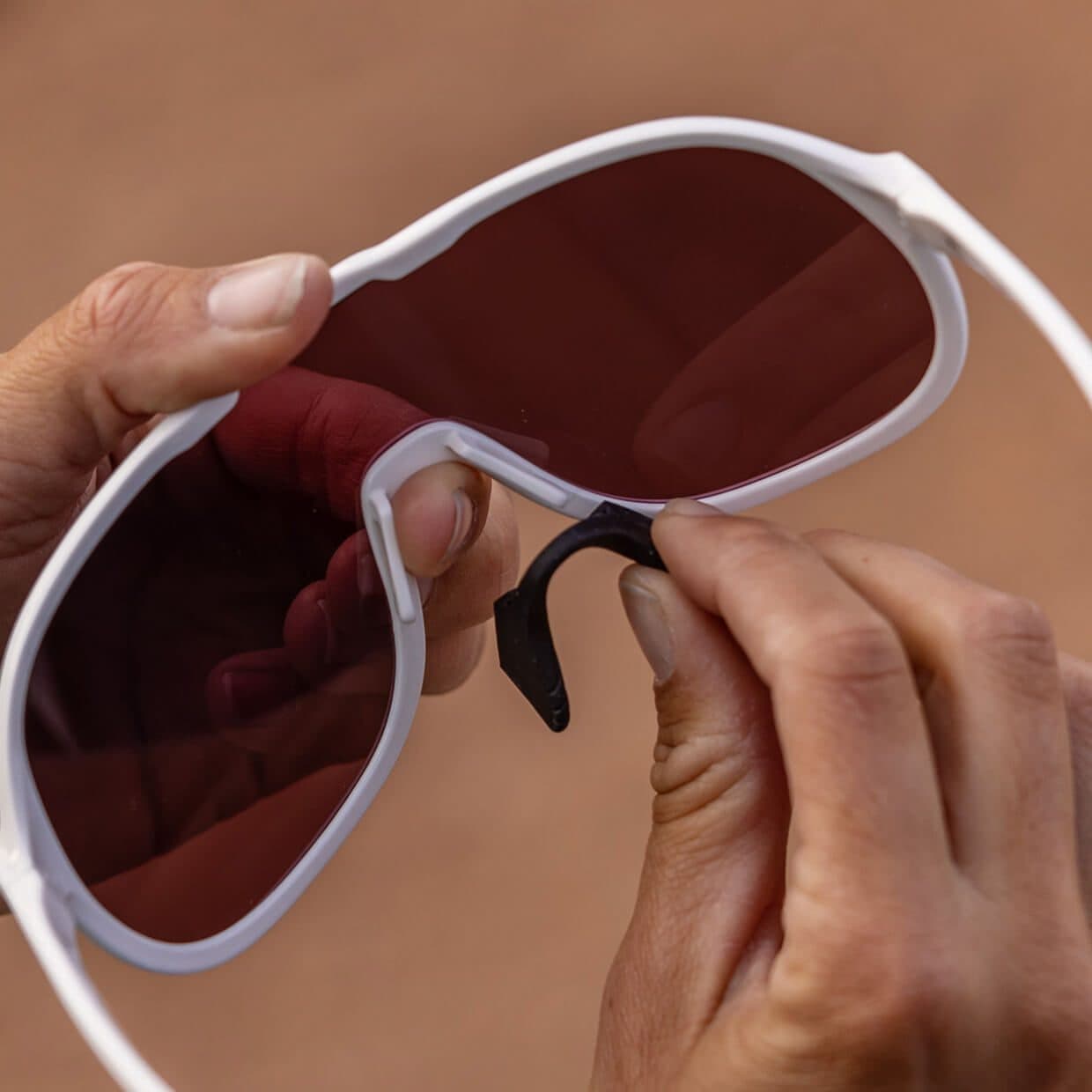 Two hands fitting a nose pad to white sunglasses with a rose lens, red rock blurred behind.