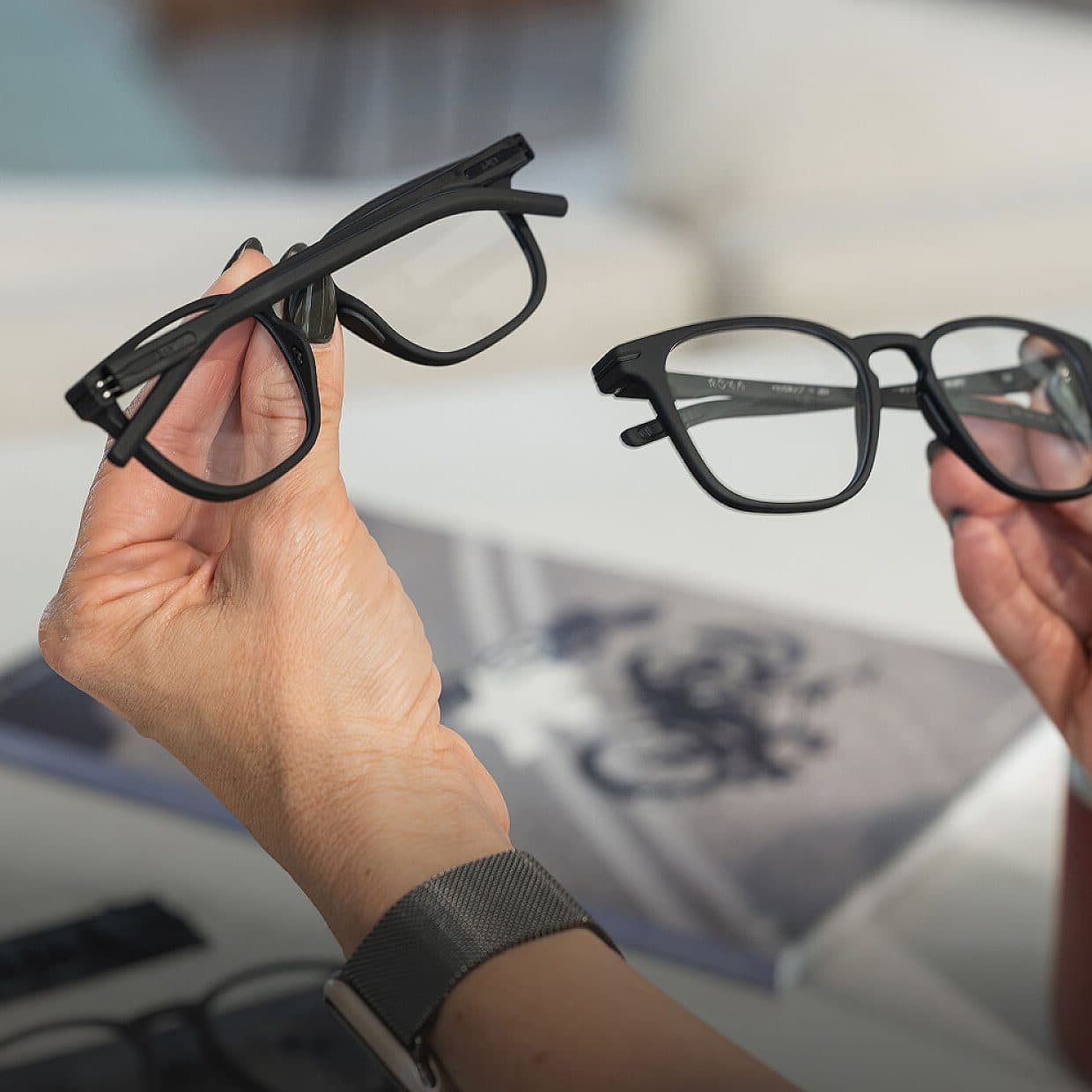 Two hands holding up two pairs of black optical frames side by side, an open try-on box and a magazine soft on the table below.