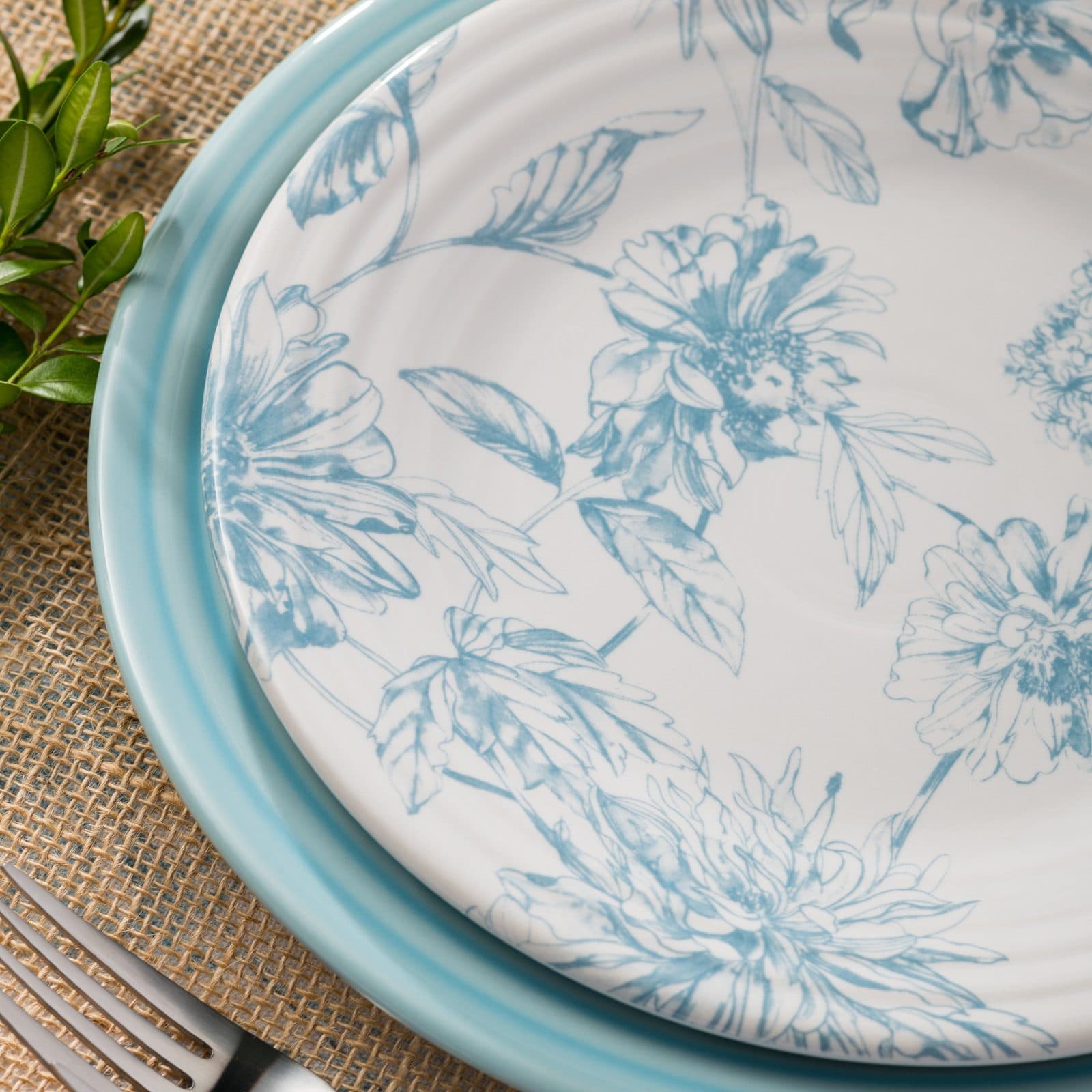 A white plate printed with blue botanical flowers, set on a turquoise plate on a burlap runner