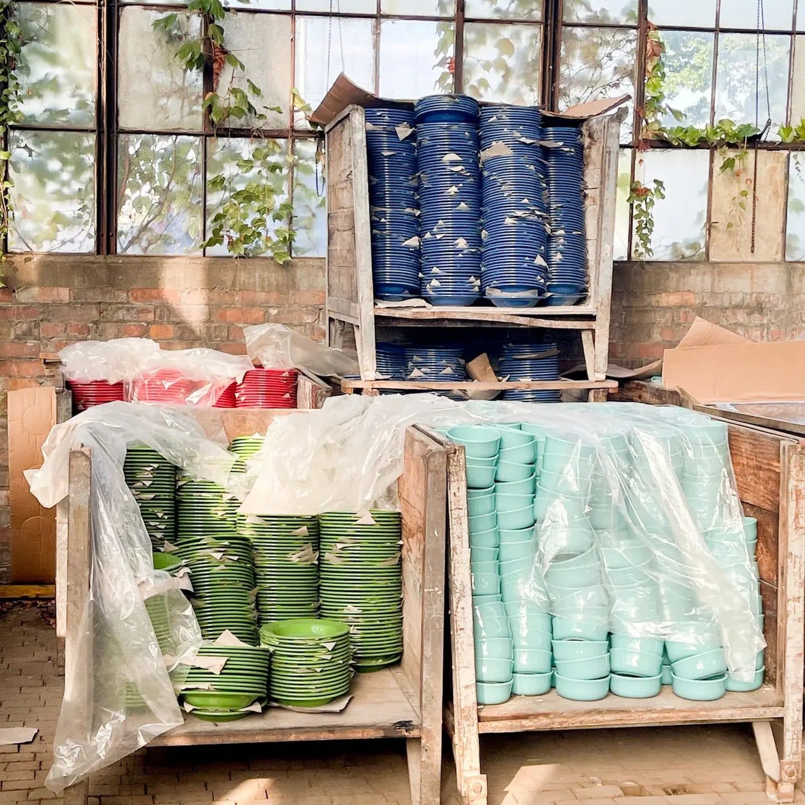 Wooden crates of cobalt, green and aqua plates half wrapped in plastic sheeting inside a brick floored factory
