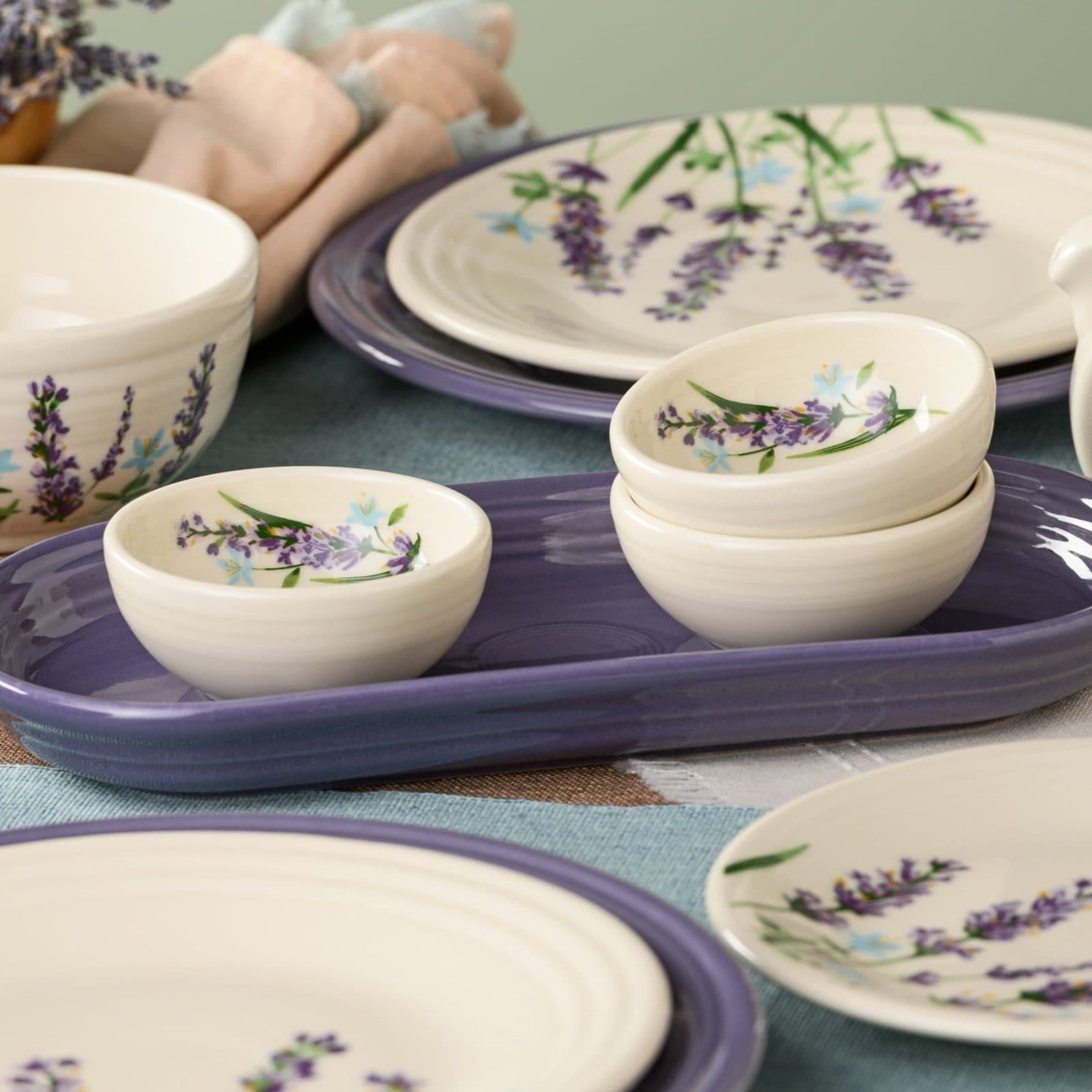 Small cream bowls painted with lavender sprigs, stacked on a lavender platter beside matching plates