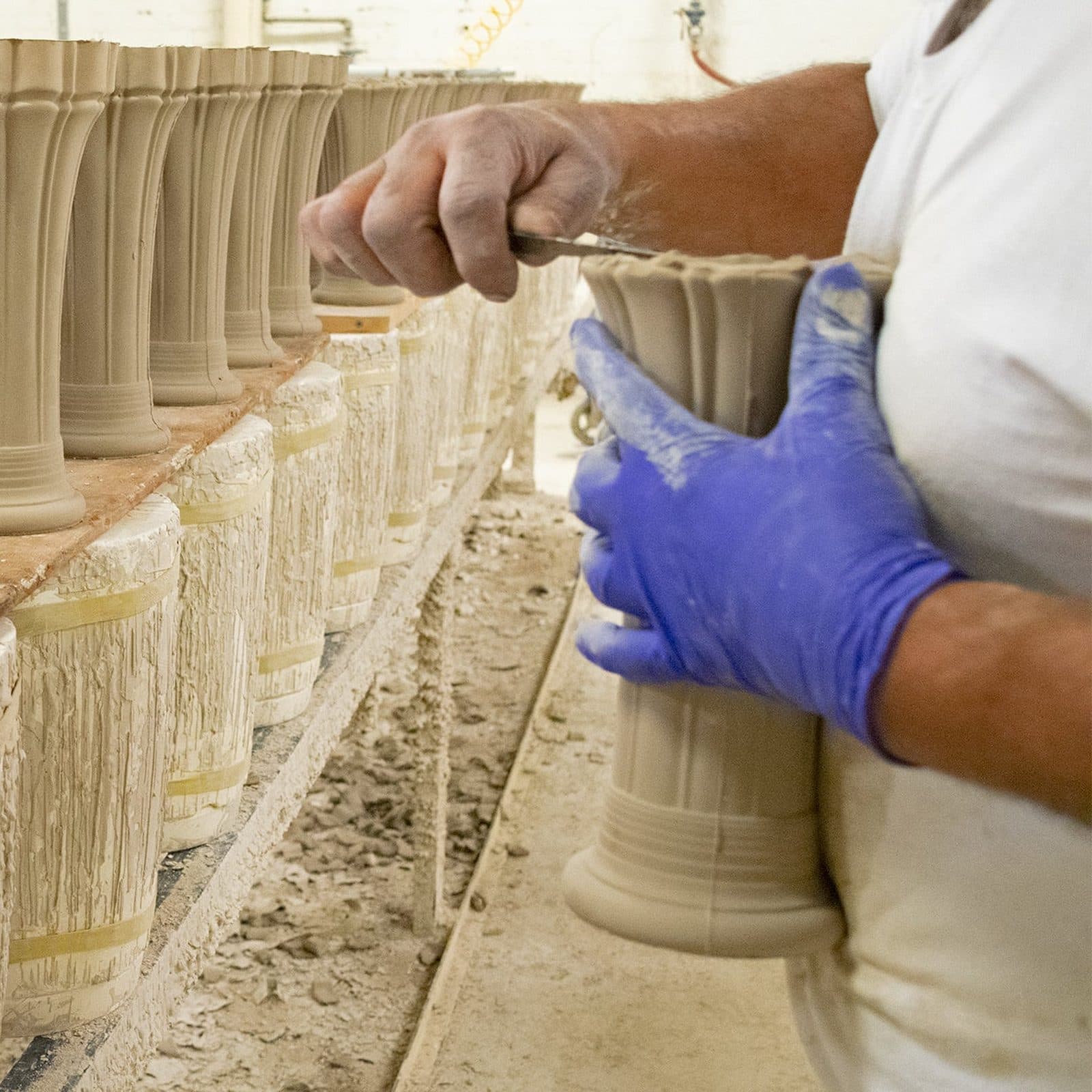A craftsman's hands, one in a blue glove, trimming the rim of a freshly cast clay vase beside a row of others