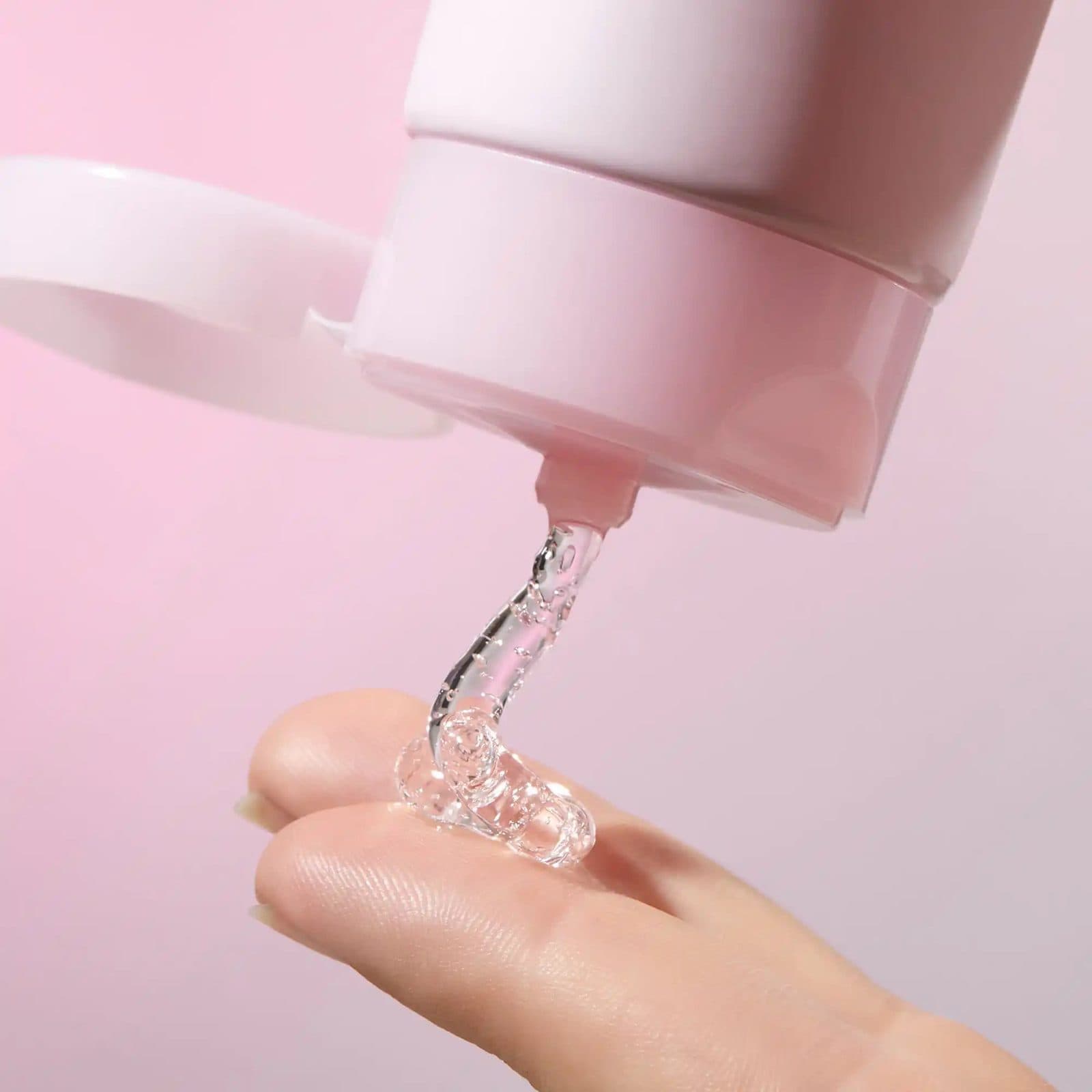 A clear gel coiling from the nozzle of a pale pink tube onto a fingertip against a pink backdrop