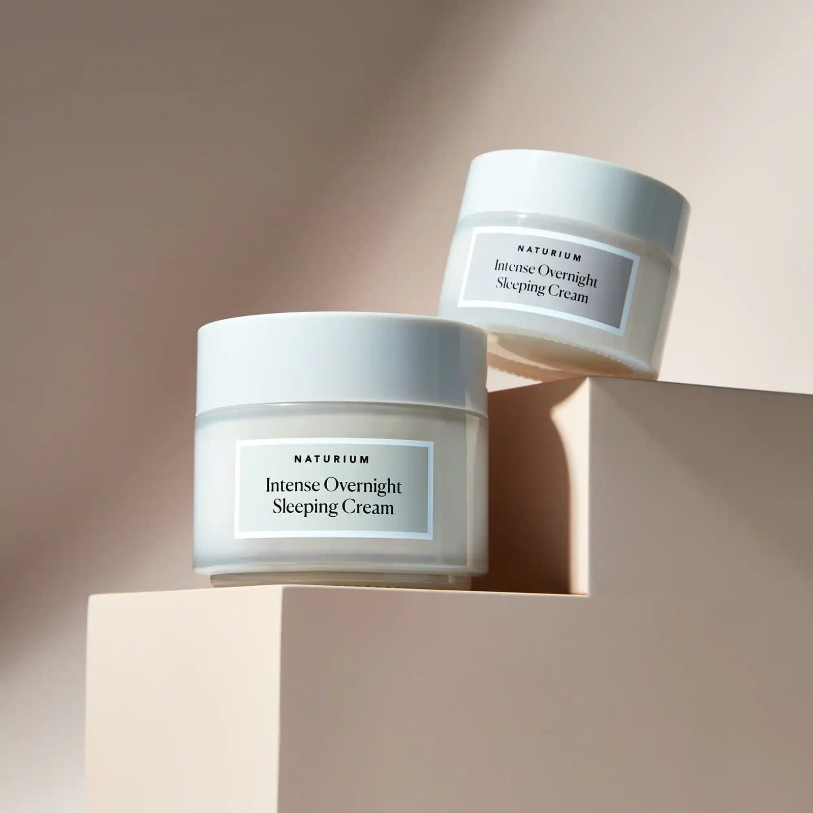 A large and a small jar of the same night cream on stepped blush plinths in soft light