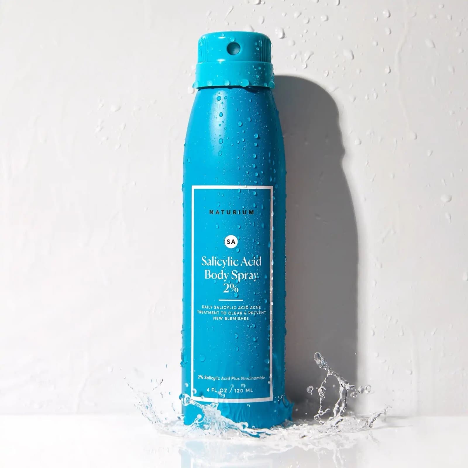 A blue body spray bottle standing in a splash of water against a white wall