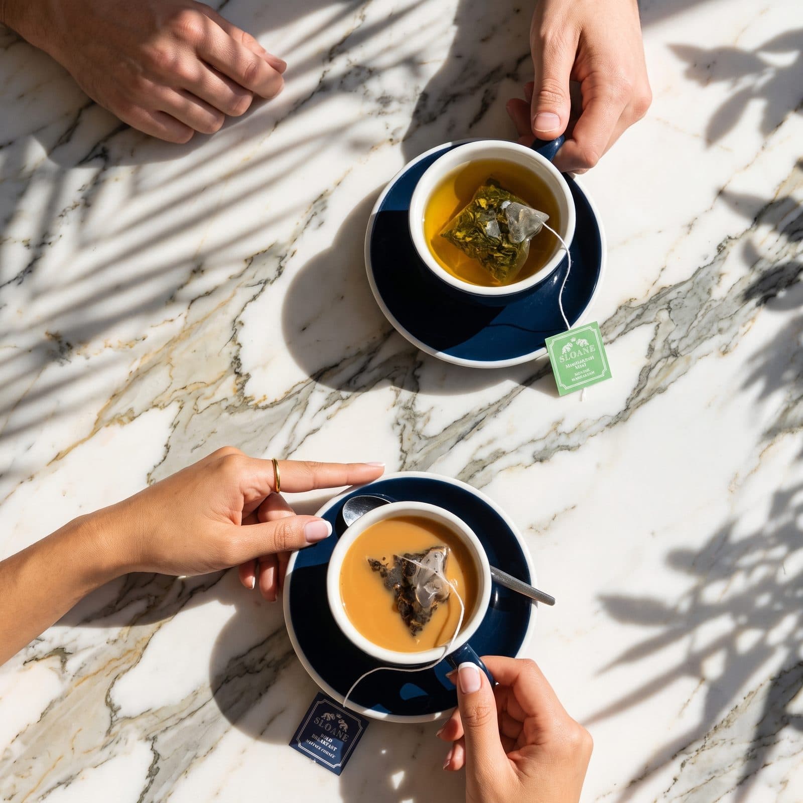 Two people's hands holding navy cups of tea across a sunlit marble table, palm leaf shadows falling over it