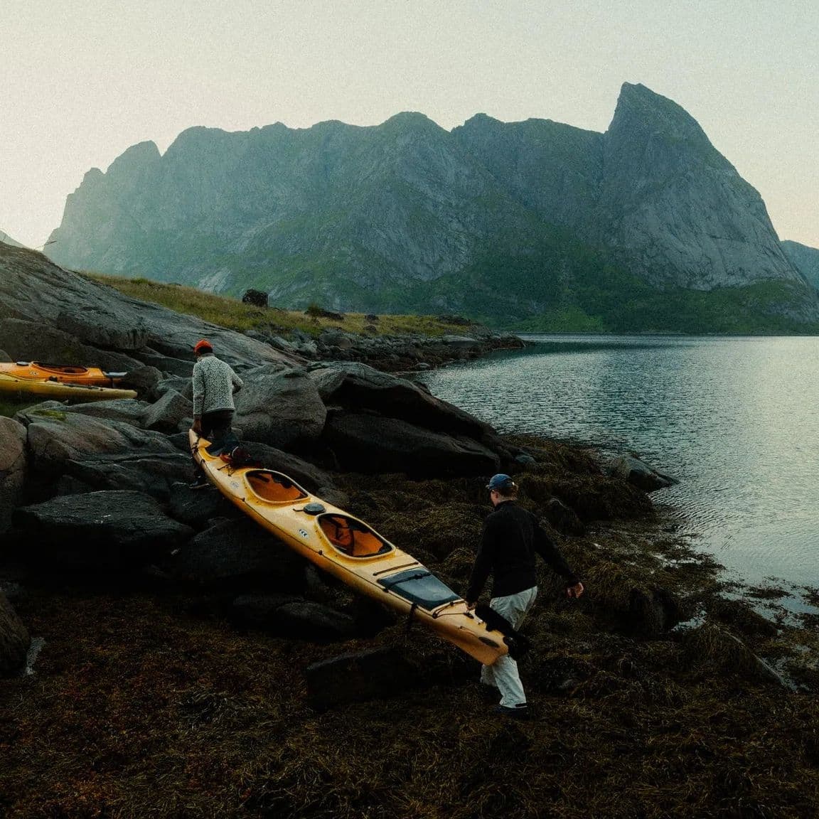 Two people carrying a yellow kayak along a rocky fjord shore at dusk, mountains behind