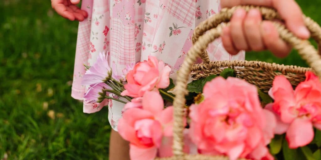 A child in a pink floral patchwork dress carrying a woven basket of pink peonies across a lawn.