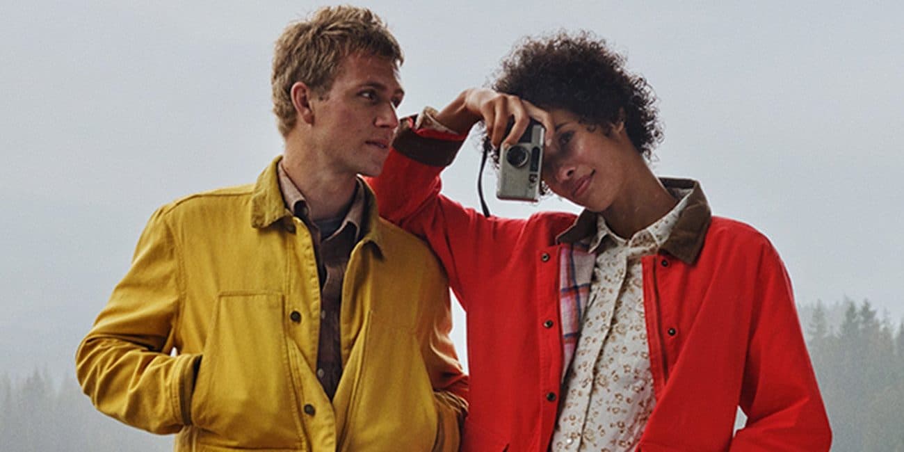 Two people by a misty lake in Eddie Bauer field jackets, one mustard yellow and one red, one holding a film camera.