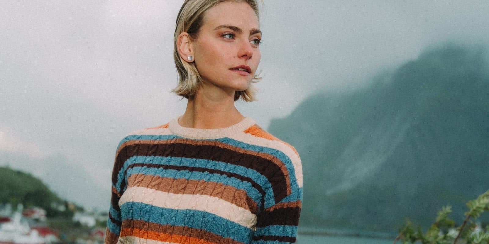 A woman with a short blonde bob in a striped cable-knit sweater, misty mountains and a fjord behind her.