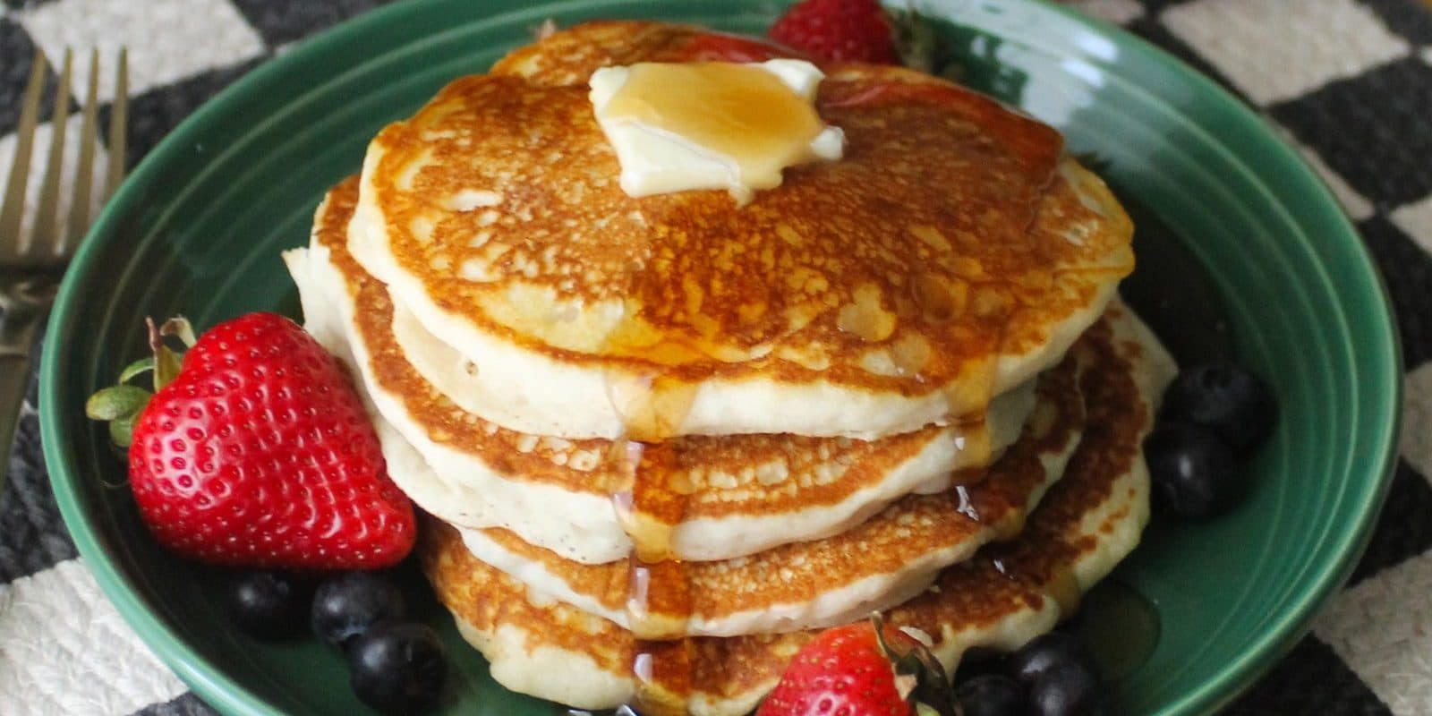 A stack of pancakes with butter, syrup, strawberries and blueberries on a jade plate over a black and white checked mat