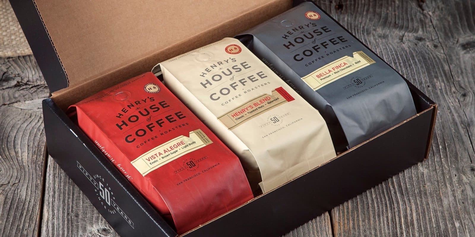 Three coffee bags in red, cream and charcoal in an open black gift box on weathered wood