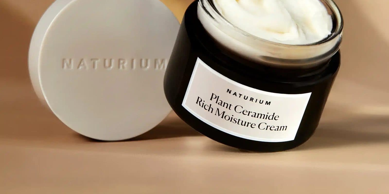 An open jar of rich moisture cream leaning beside its embossed lid on a warm tan backdrop