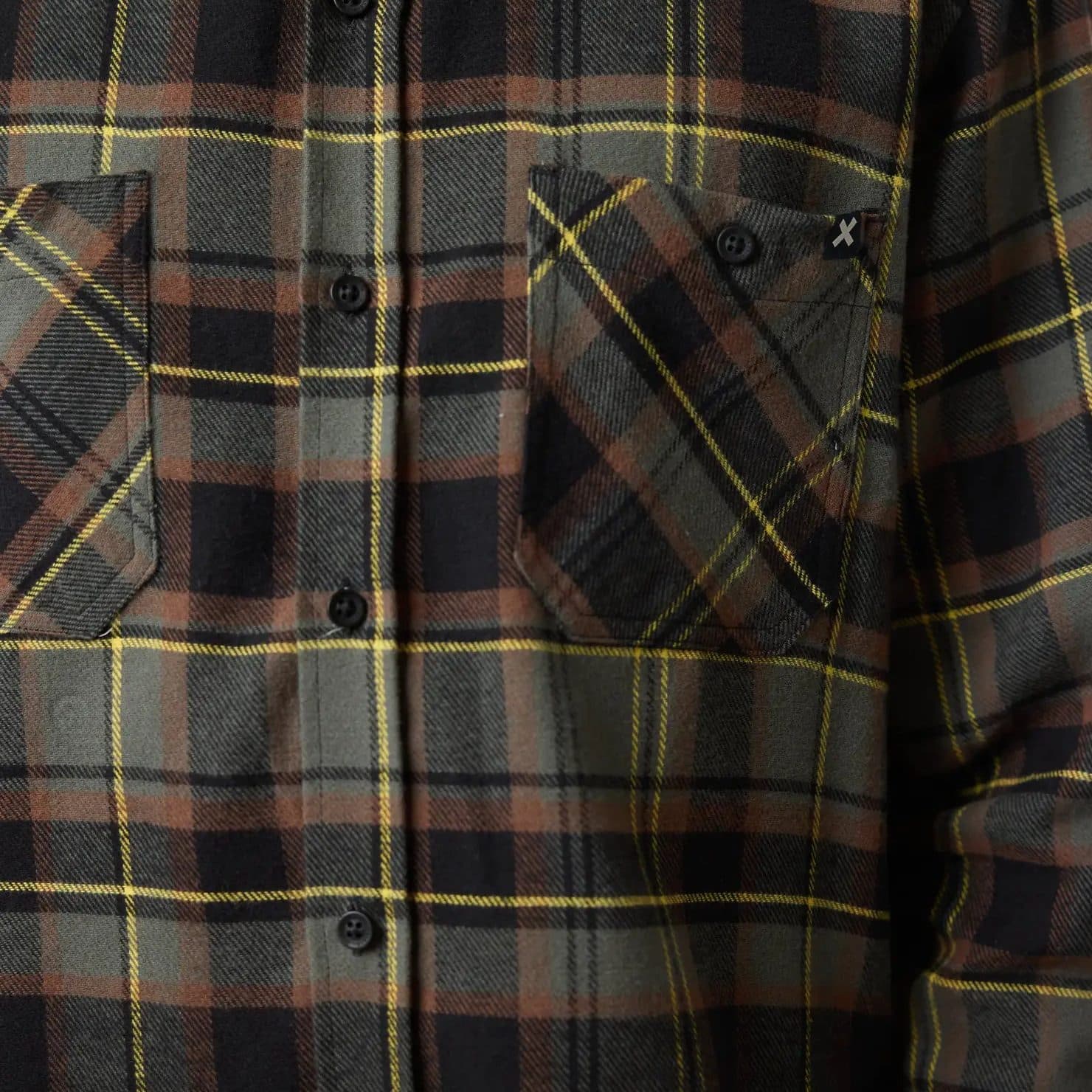 A buttoned plaid flannel shirt in olive, rust and black, close up at the chest pocket.