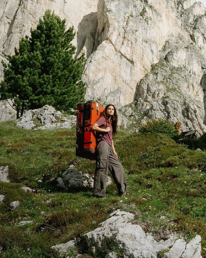 A hiker in a maroon Blue Banana tee carrying an orange pack across a green alpine slope below limestone cliffs.