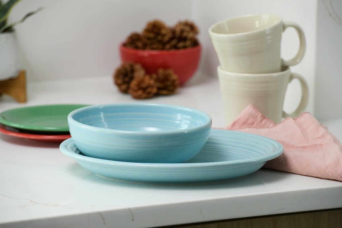 A turquoise Fiesta bowl and plate with cream Fiesta mugs and a pink napkin on a kitchen counter.
