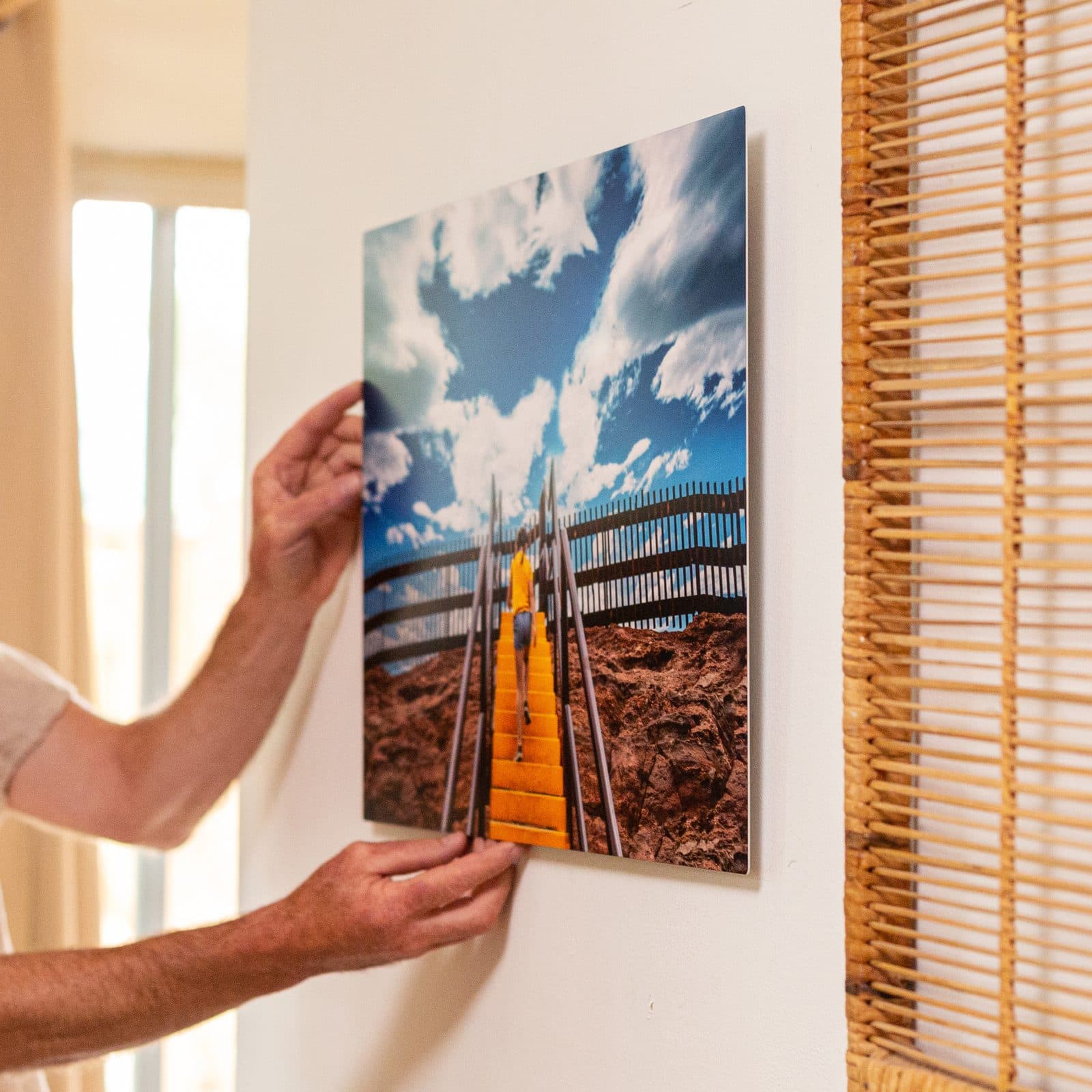 Two hands pressing a framed print of a figure climbing a yellow stair against a cream wall beside a rattan blind