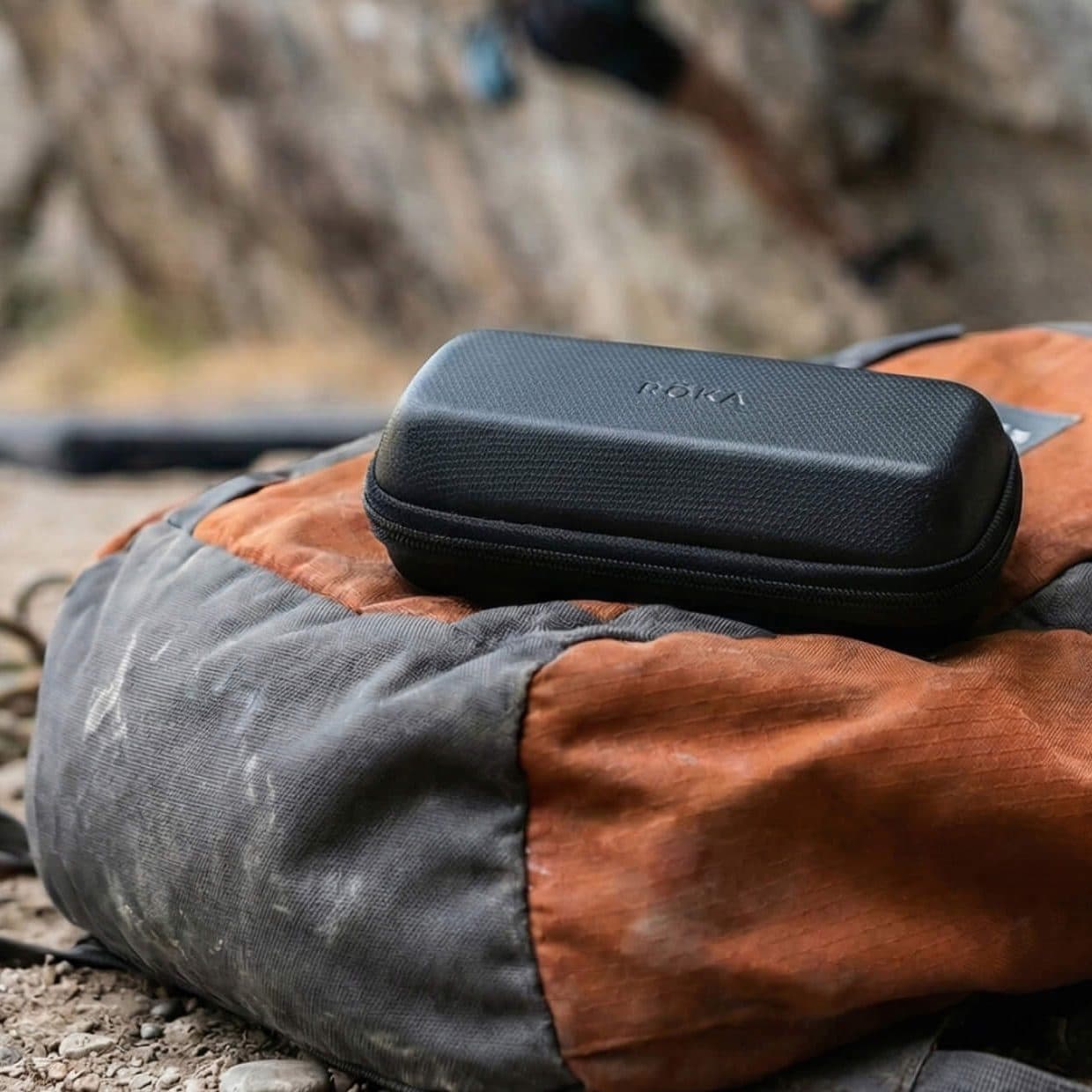 A black glasses case resting on a dusty orange and grey backpack at the foot of a climbing wall, a rope coiled behind.